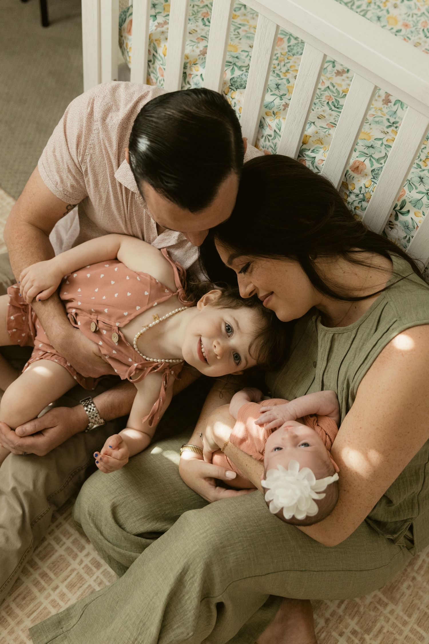 A family of four sits by a white crib, parents lovingly holding their toddler and newborn baby in a cozy nursery.