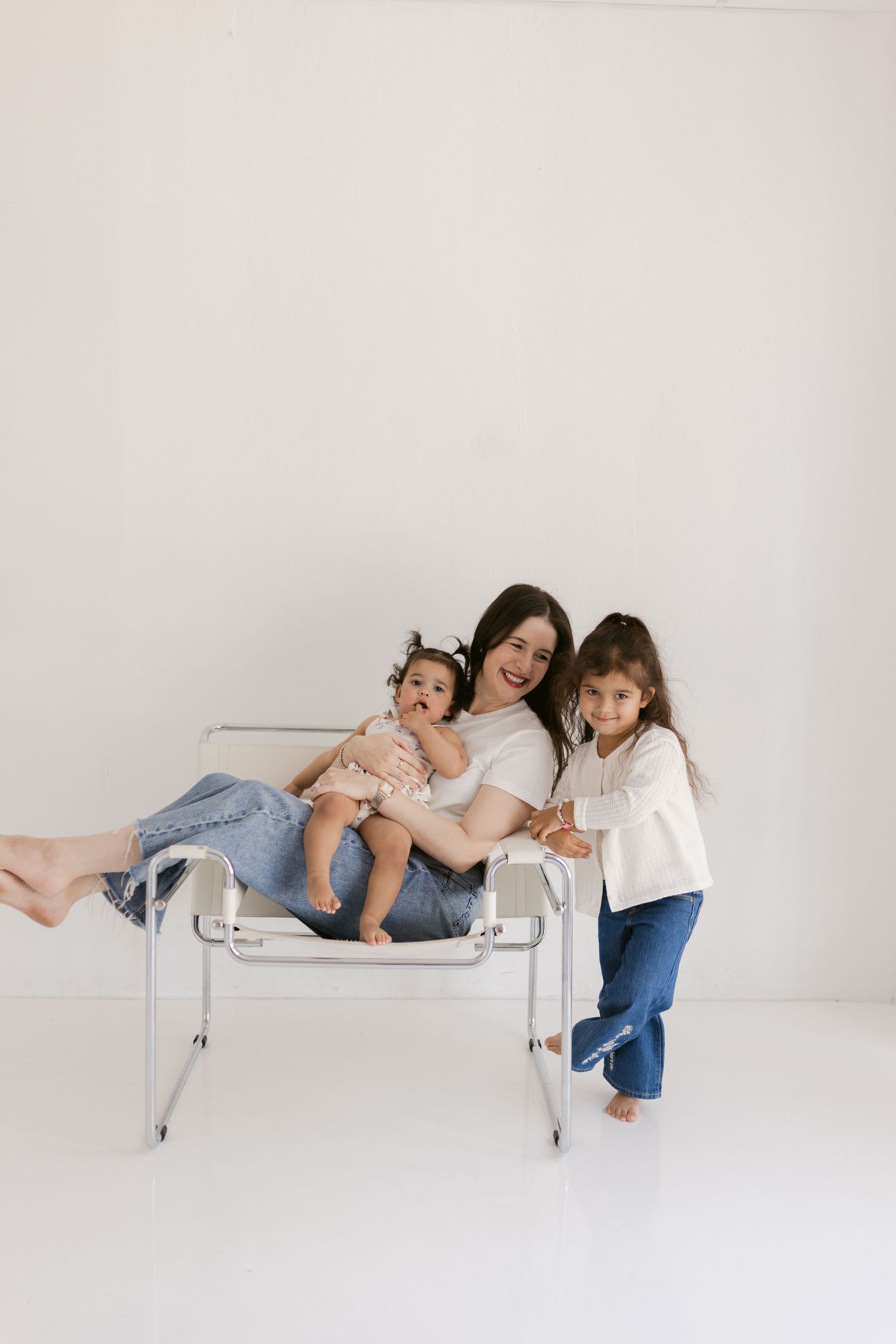 A mother sits on a modern chair holding her baby while her young daughter leans in smiling, in a bright white studio.
