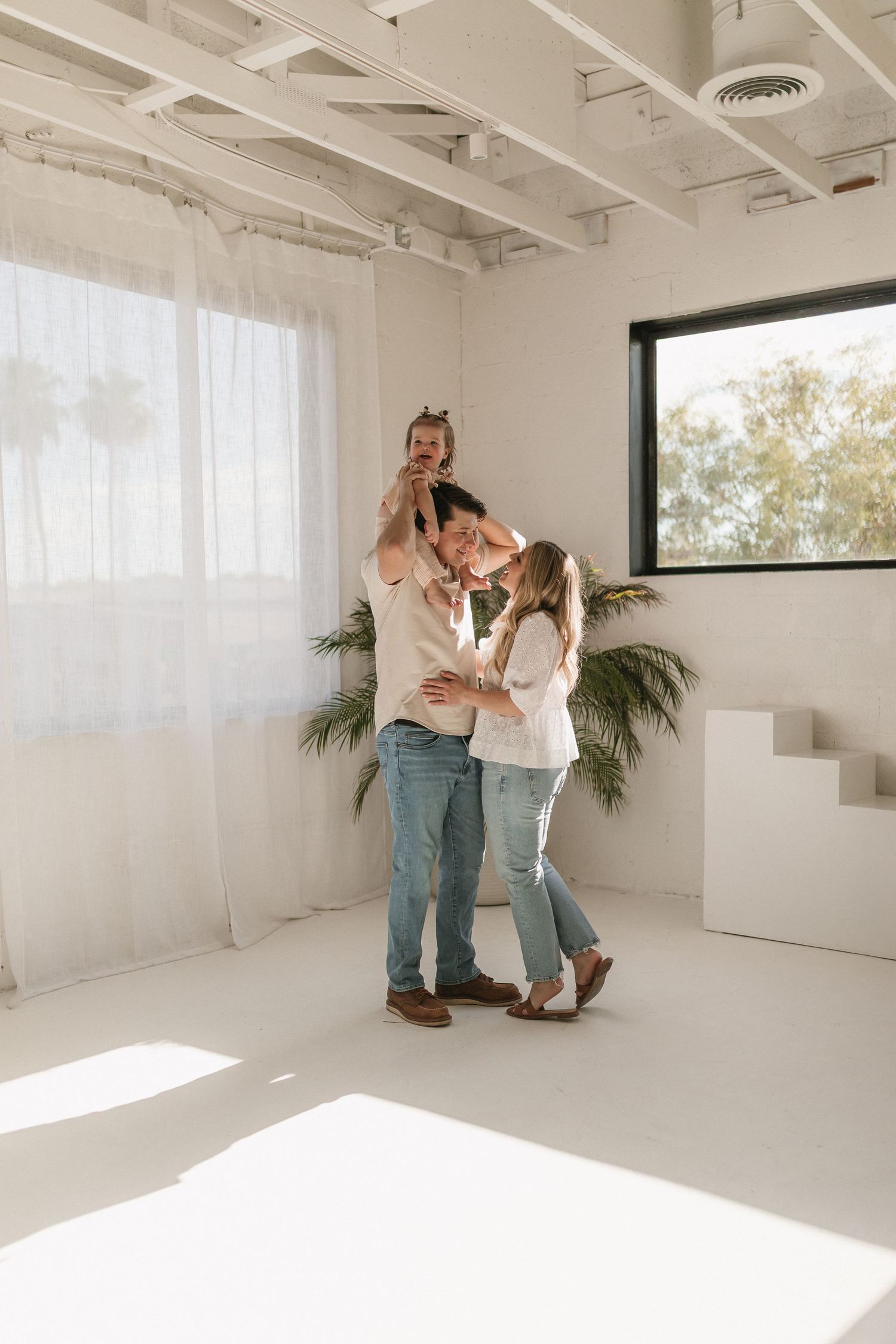 A couple shares an intimate moment together in a bright white room with palm plants and natural sunlight streaming in.