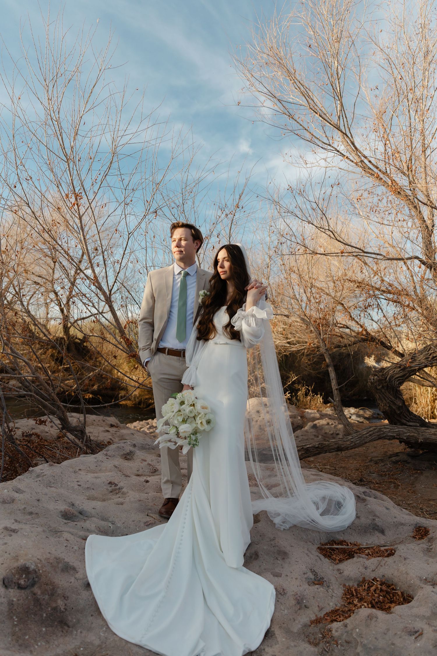 A couple in wedding attire poses together on a sandy landscape with bare winter trees in the background.