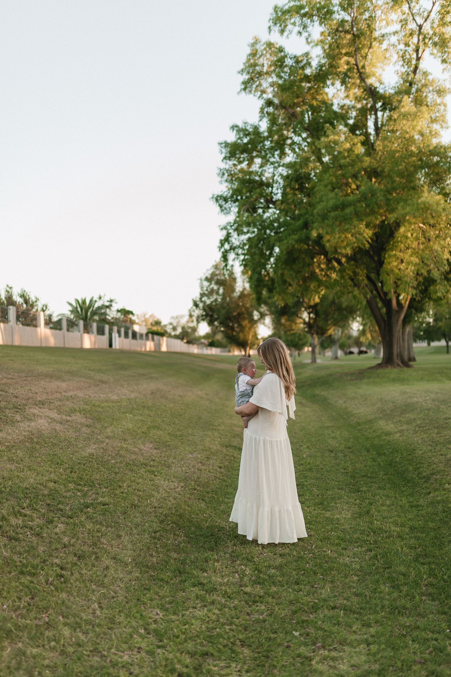 Woman in a white dress stands in a grassy park setting during golden hour with trees in the background.