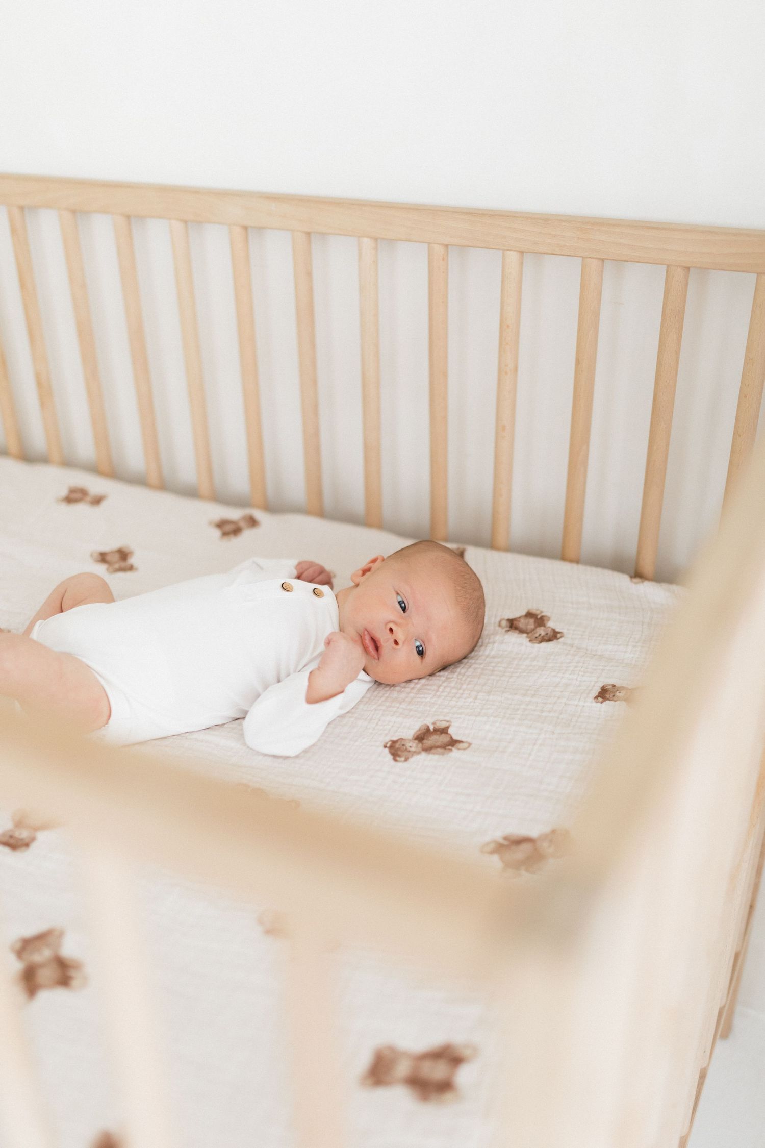 A baby lies peacefully in a white wooden crib with teddy bear print bedding.