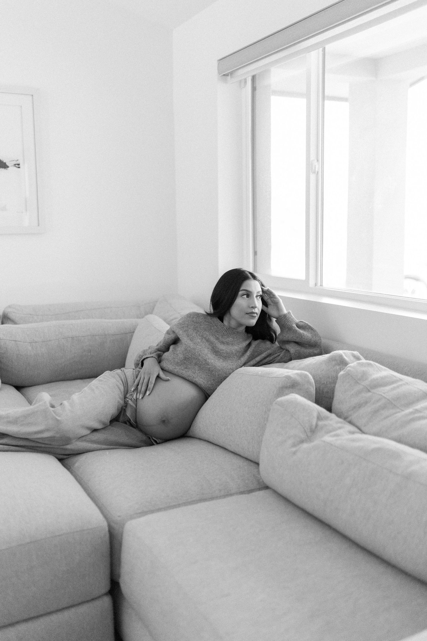 Black and white photo of a pregnant woman relaxing on a sectional sofa, gazing out a bright window.