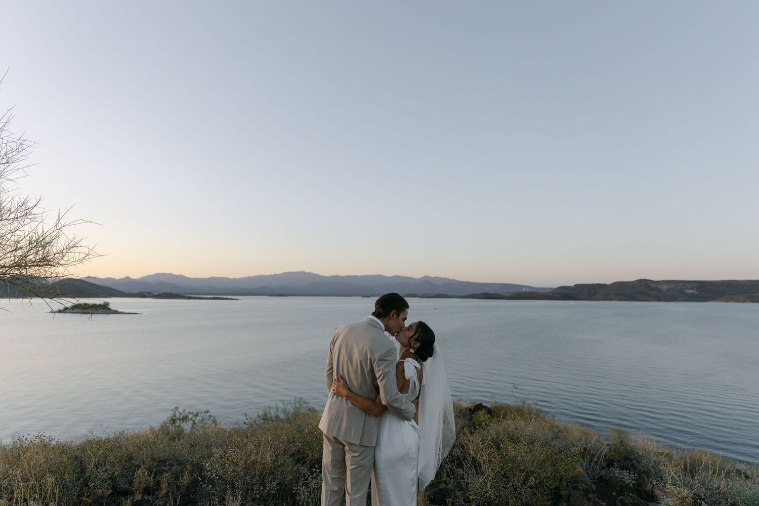 A couple in wedding attire stands at the edge of a lake during golden hour.