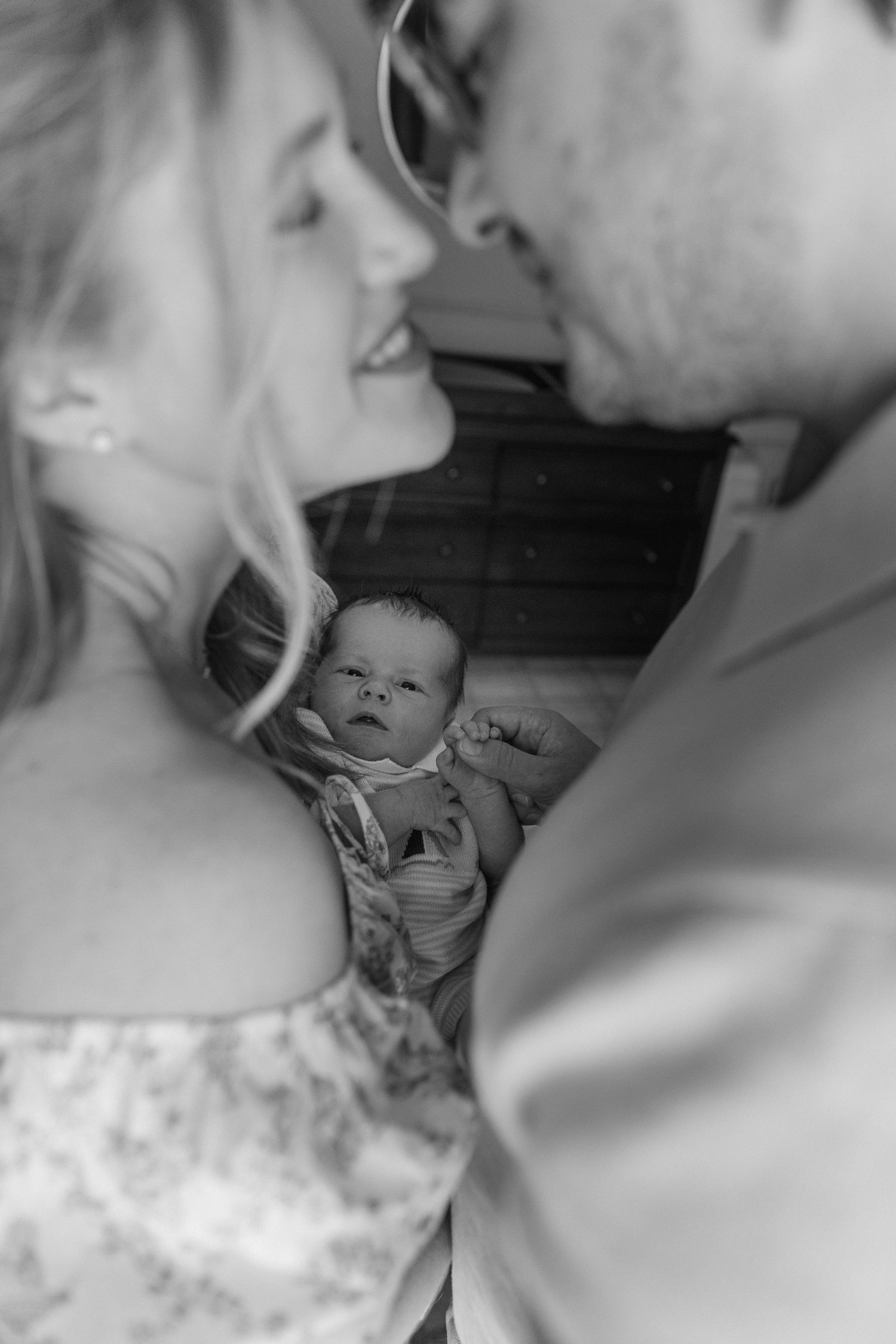 Black and white portrait of parents leaning close together with their newborn baby nestled between them.