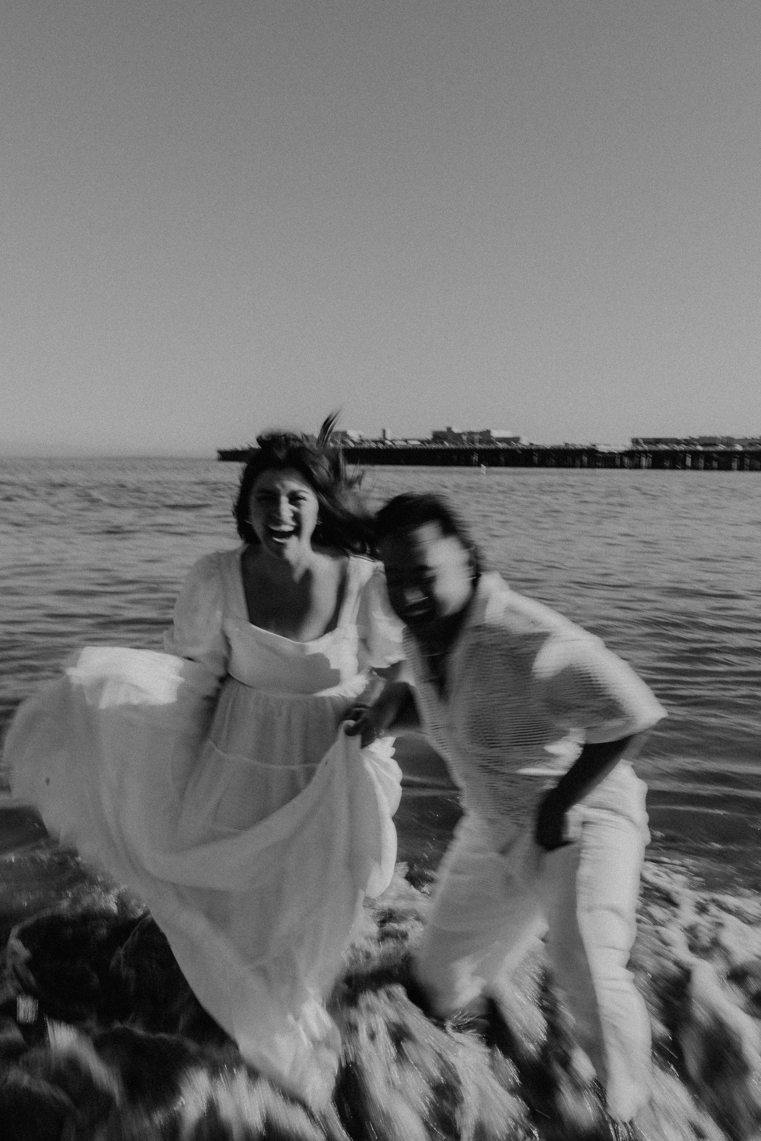 Black and white photo of couple playing in ocean waves wearing white outfits.