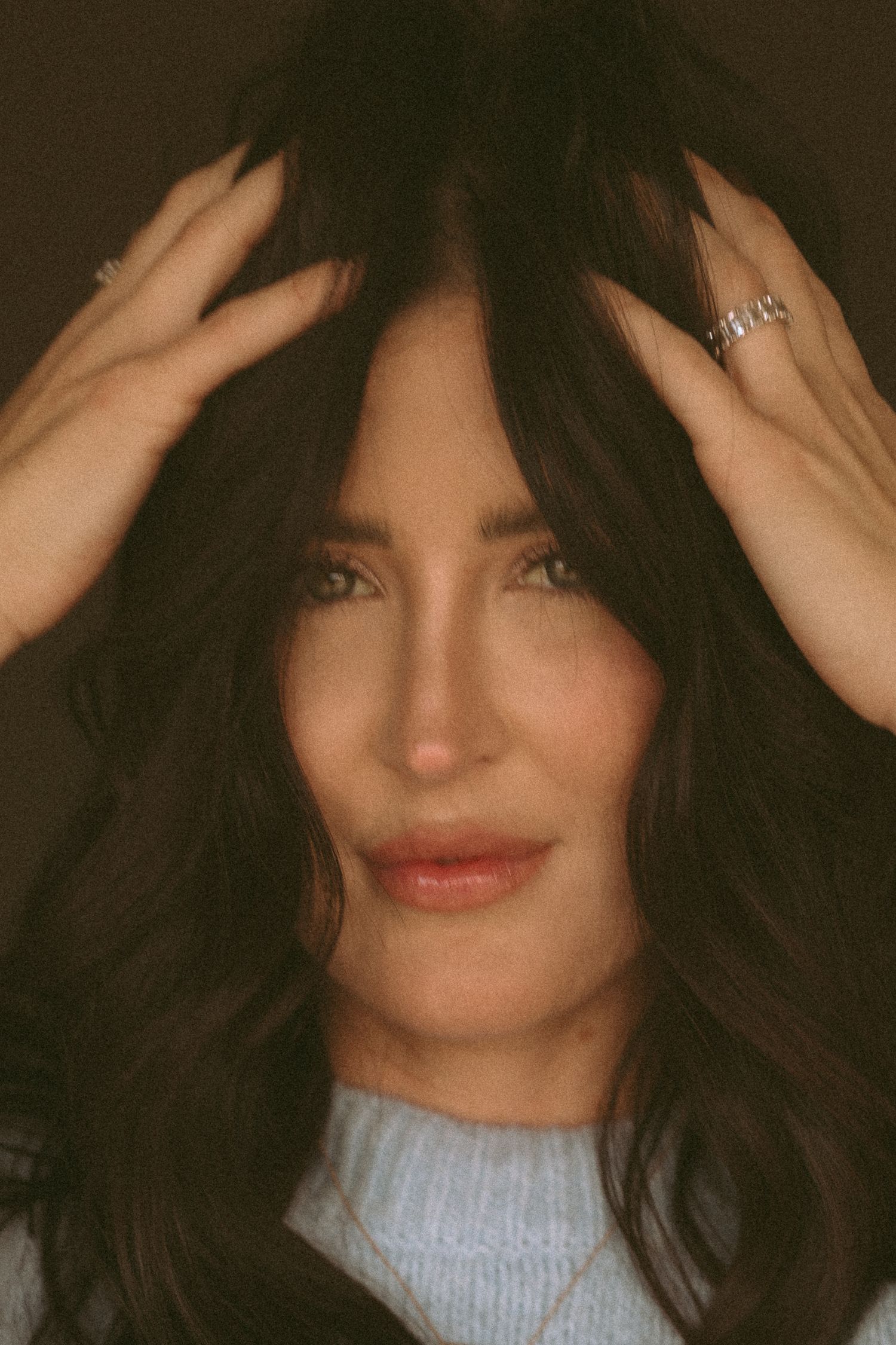 A close-up portrait of someone with long dark wavy hair wearing a light blue sweater in warm sepia lighting.