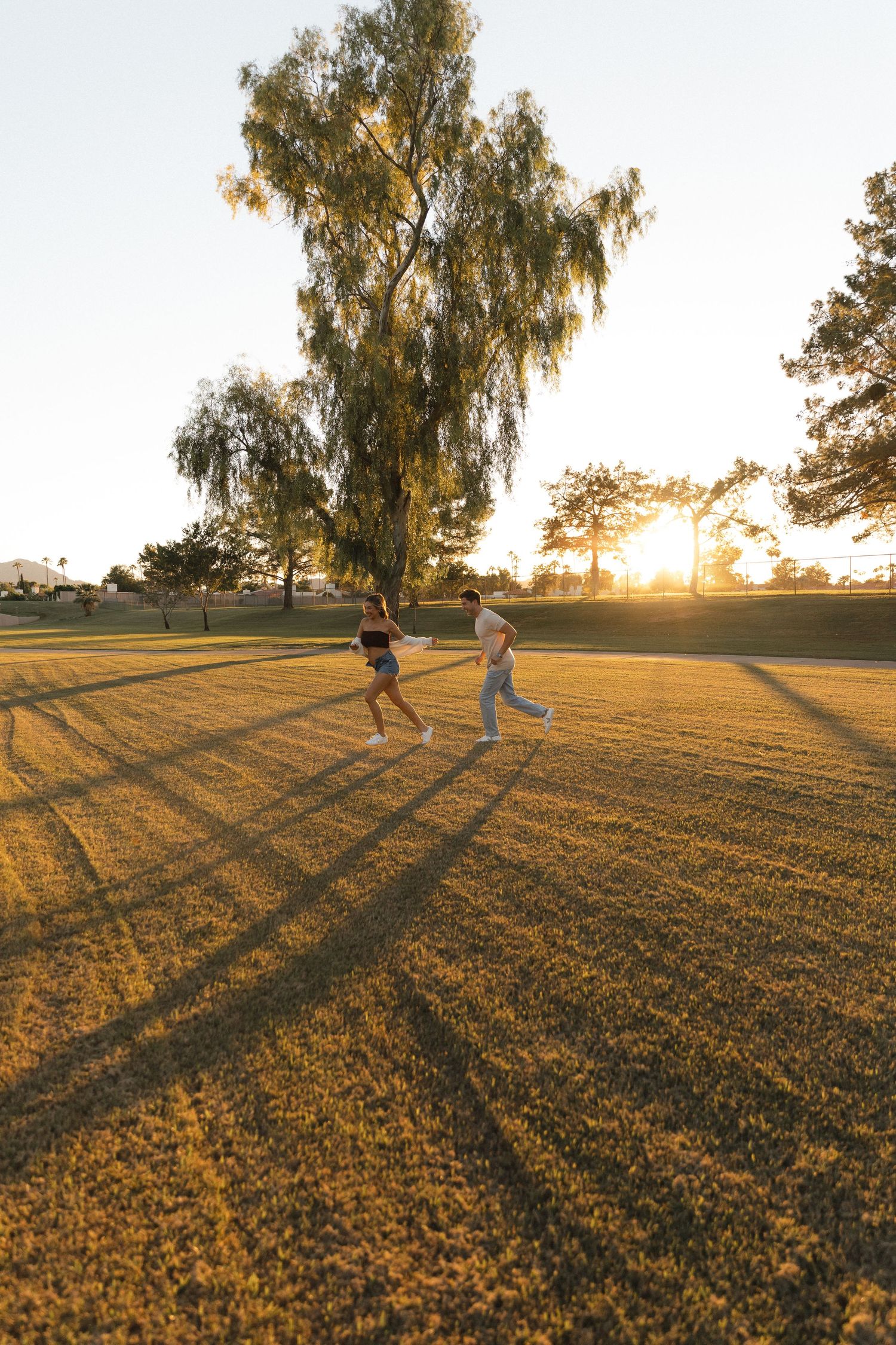 Two people running across a grassy field at sunset with long shadows cast by trees in the golden light.