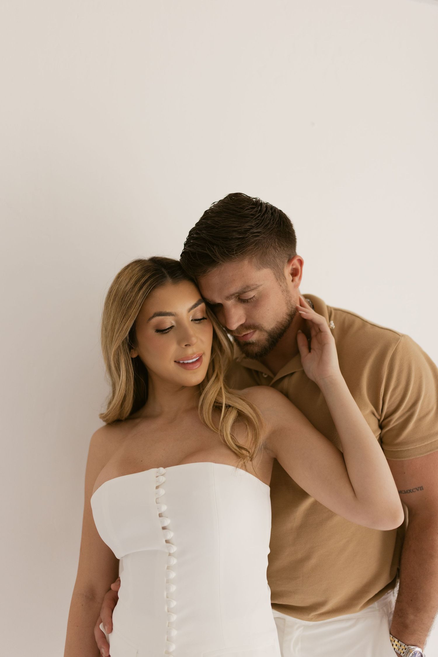 A romantic couple poses together, woman in white strapless dress, man in tan shirt, against a white background.
