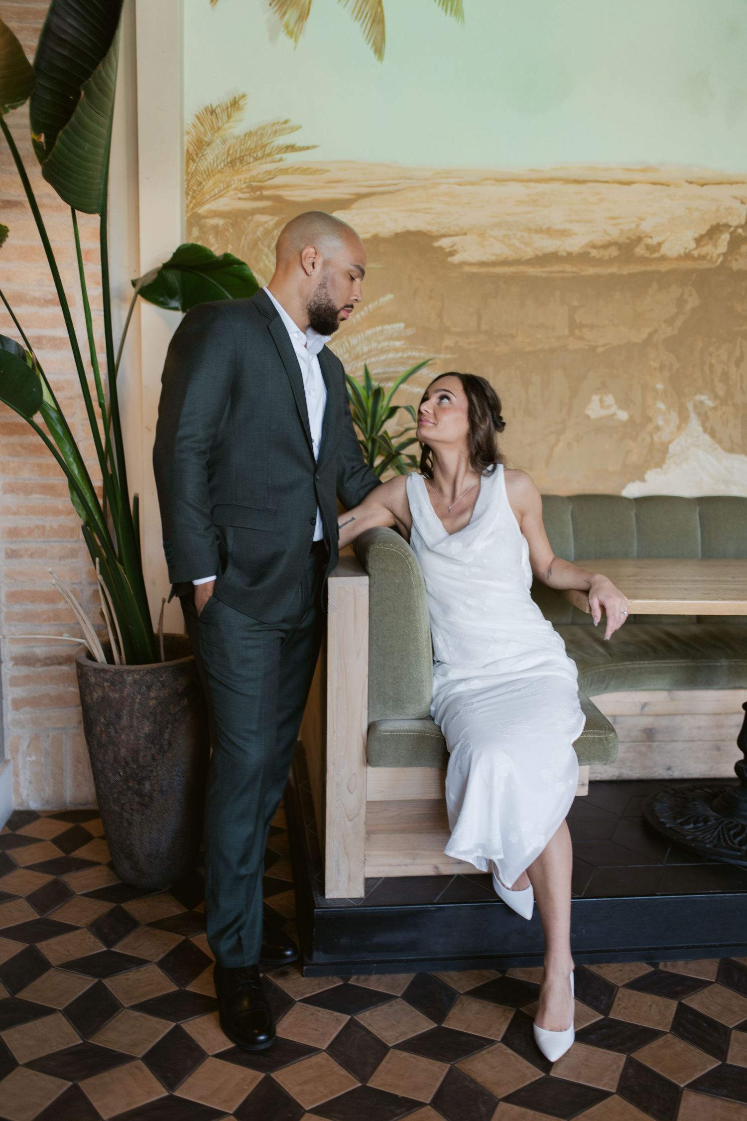A stylish couple poses casually against a wall with tropical plants and checkered flooring.