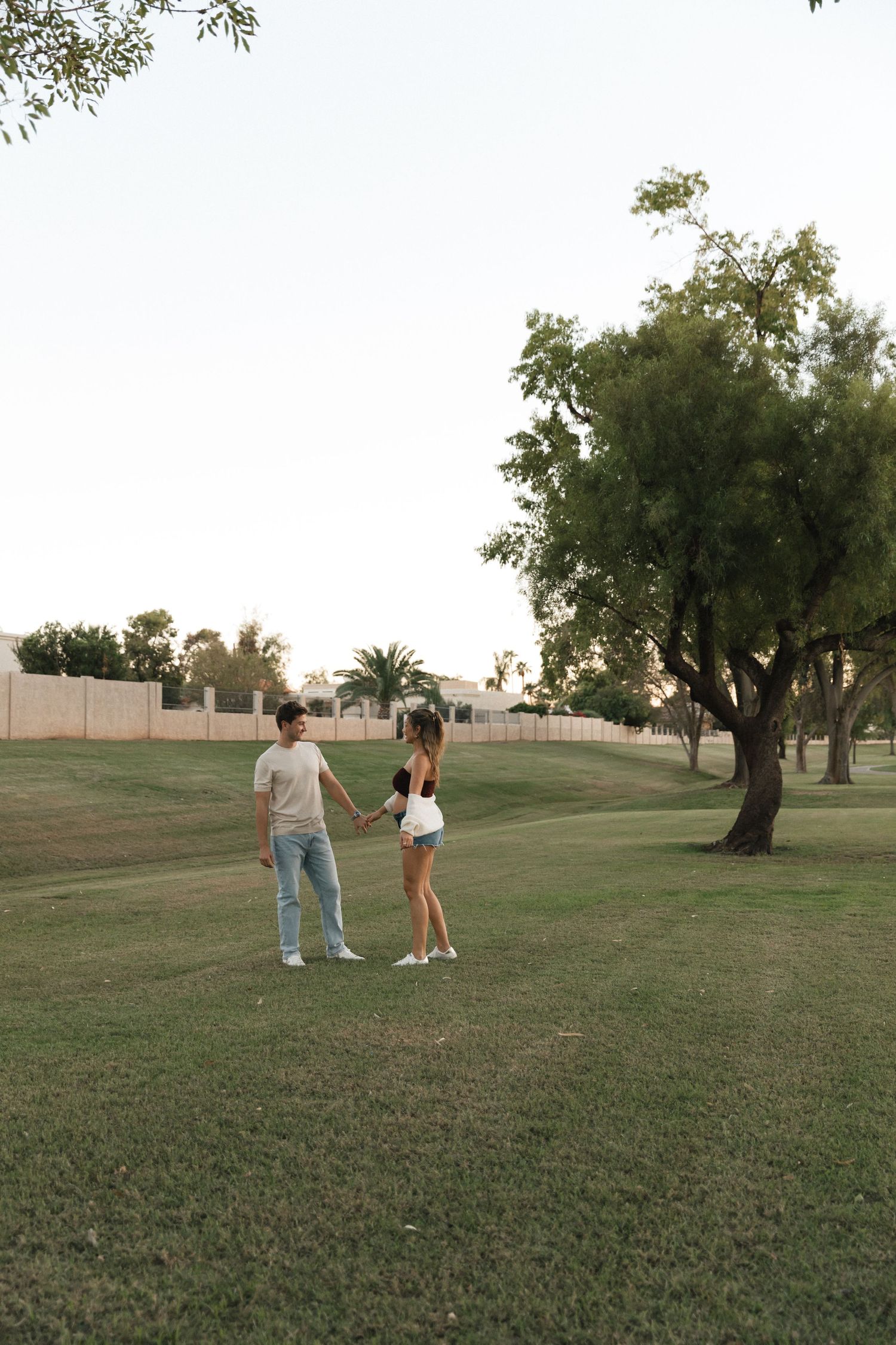Two people stand together on a grassy hill at sunset with trees scattered in the background.