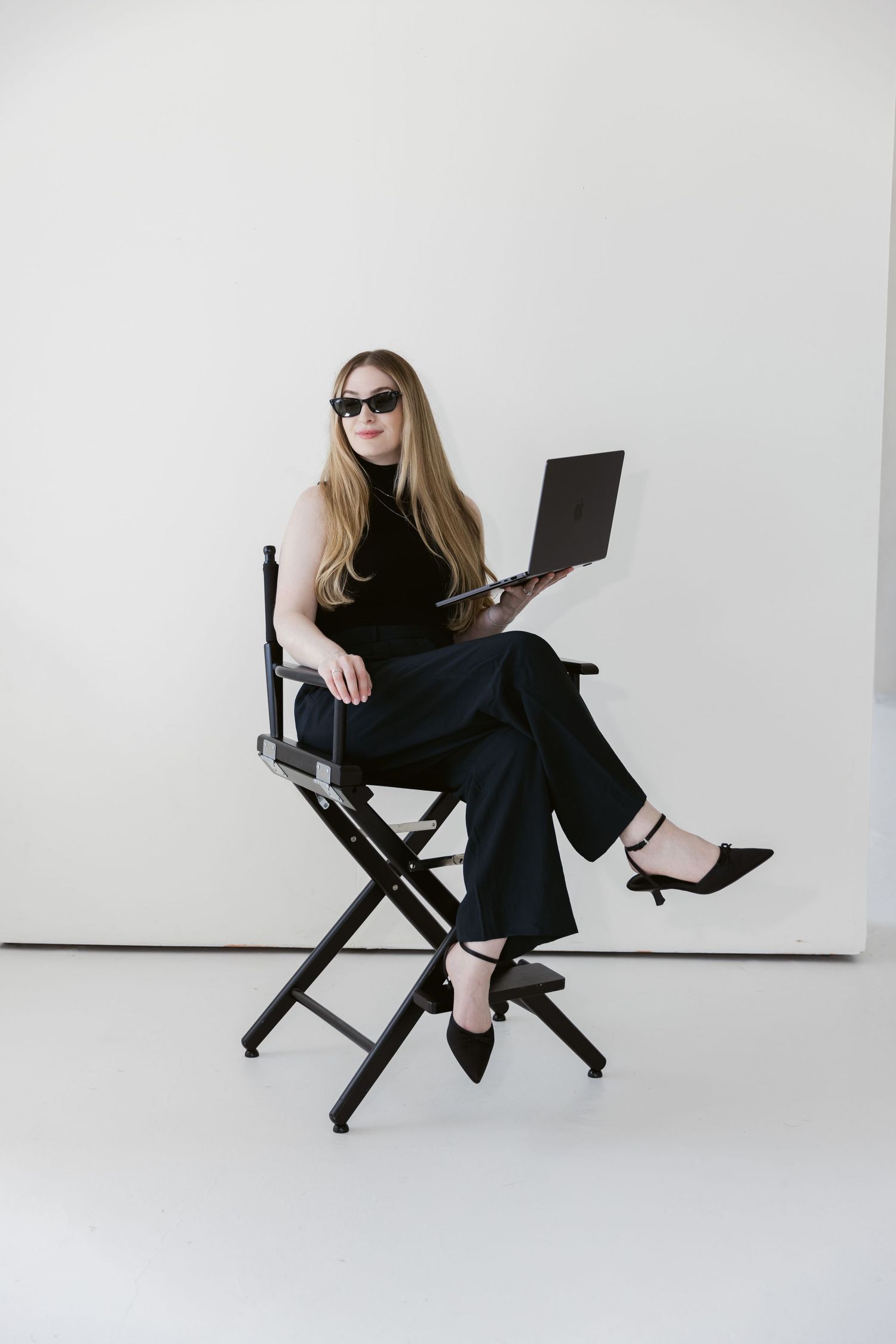 Woman in black outfit and sunglasses sitting in director's chair holding laptop against white background.