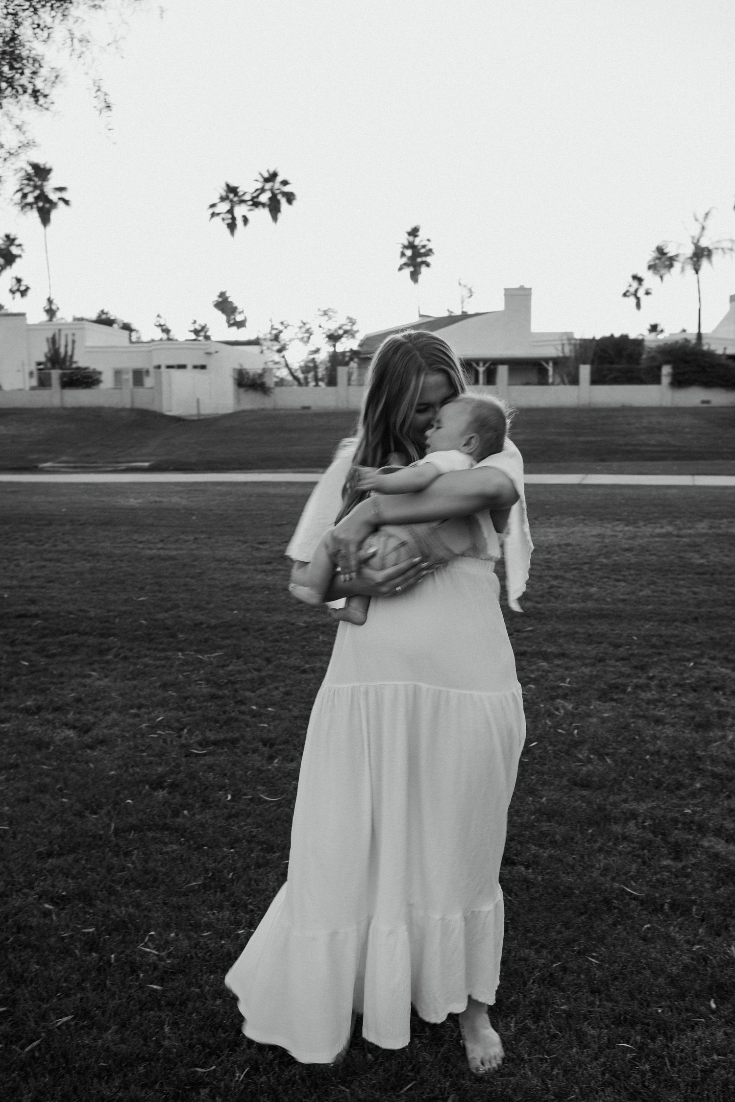Three people in white dresses embrace while walking across a grassy field as sunset approaches.