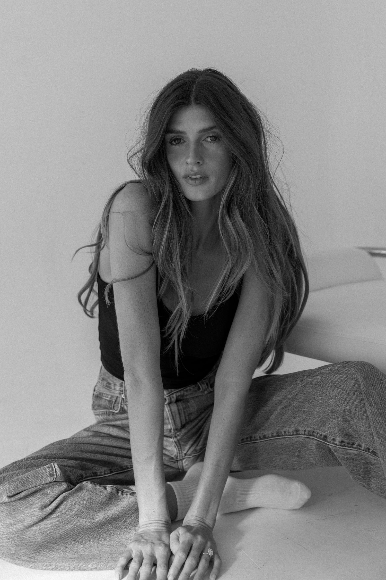 Black and white photo of a young woman with long hair sitting casually in jeans and a black tank top.