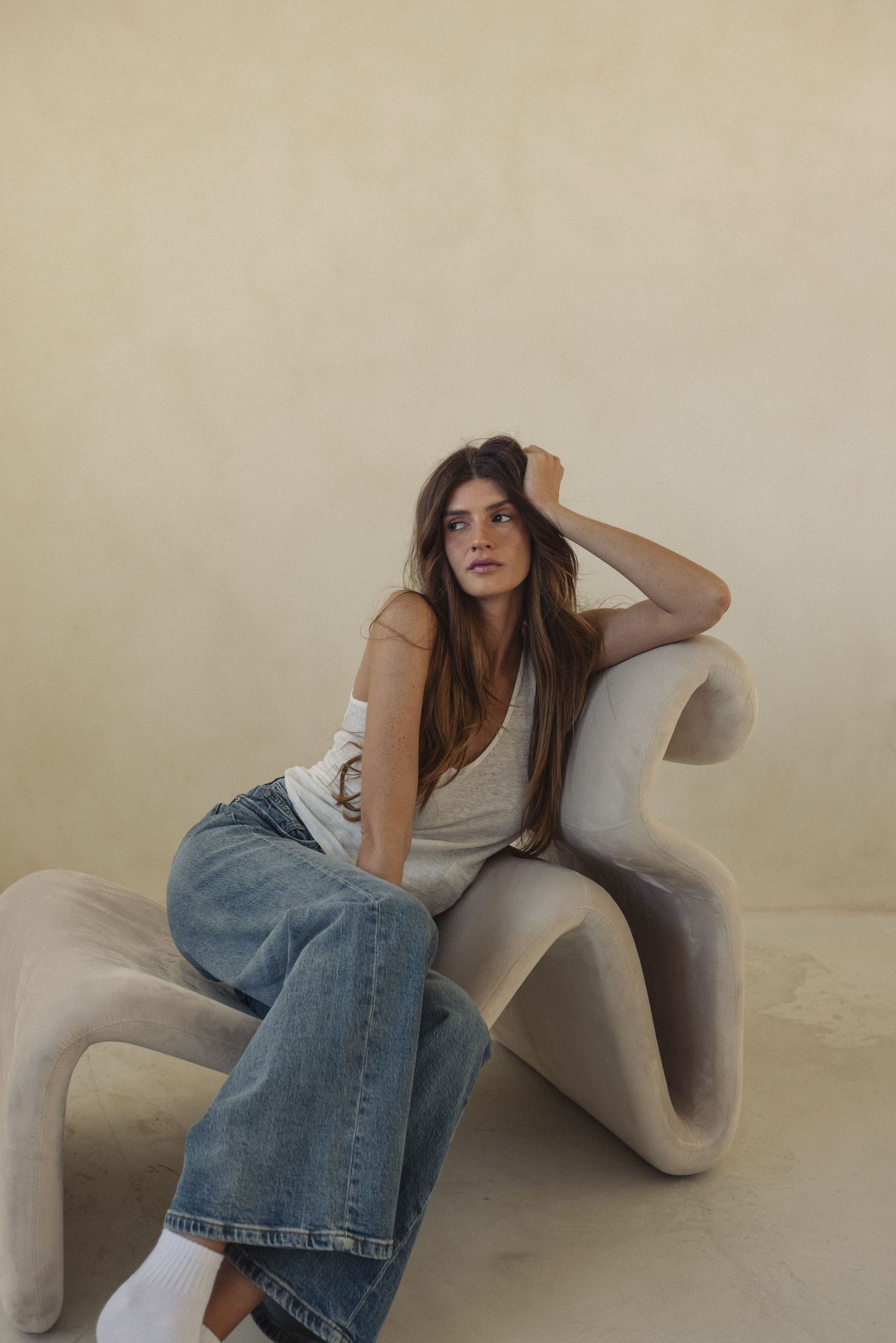 Young woman in white tank top and wide-leg jeans sitting in a sculptural beige chair against a neutral wall.