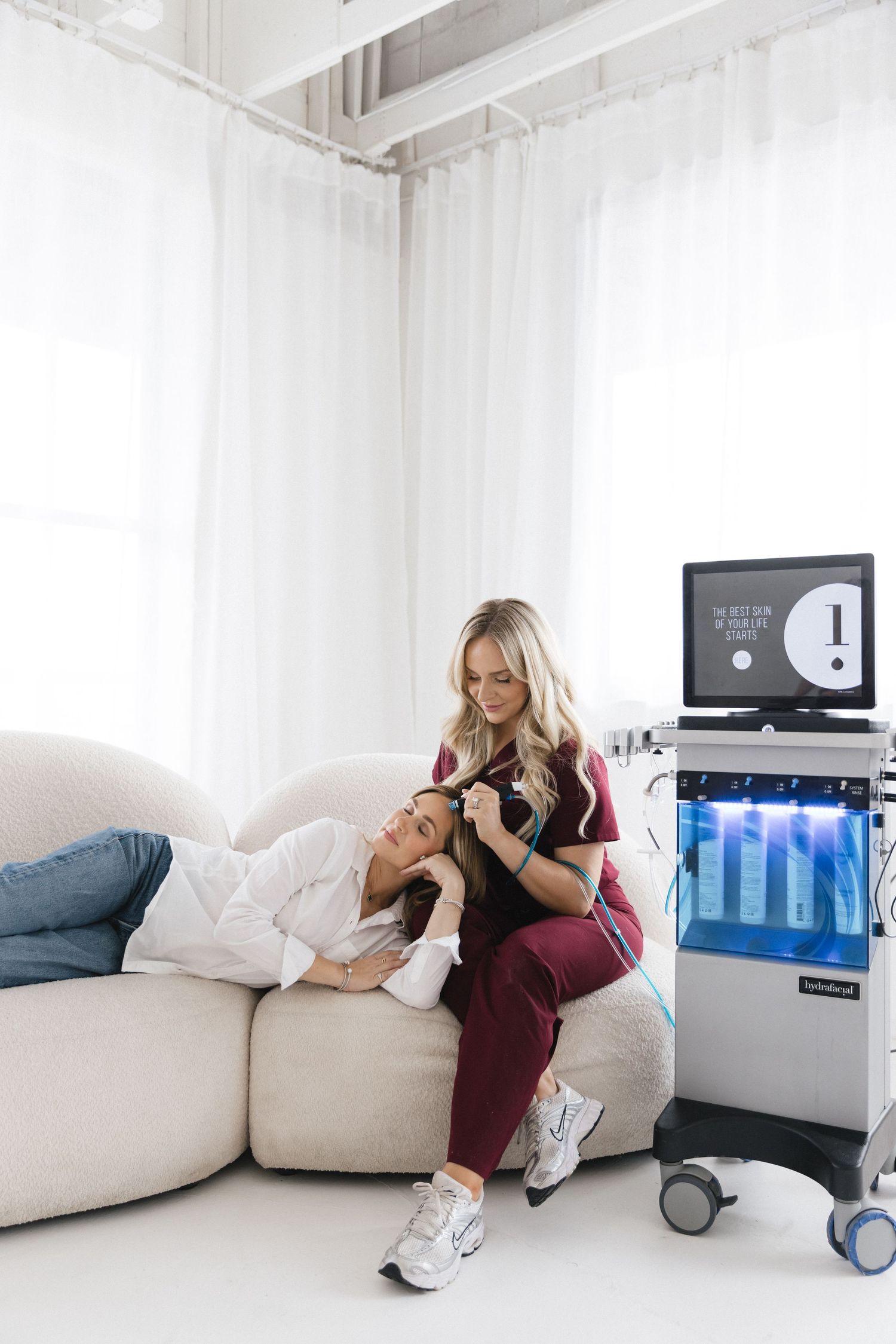 A blonde aesthetician in burgundy scrubs consults with a client on a white sofa beside a HydraFacial machine.