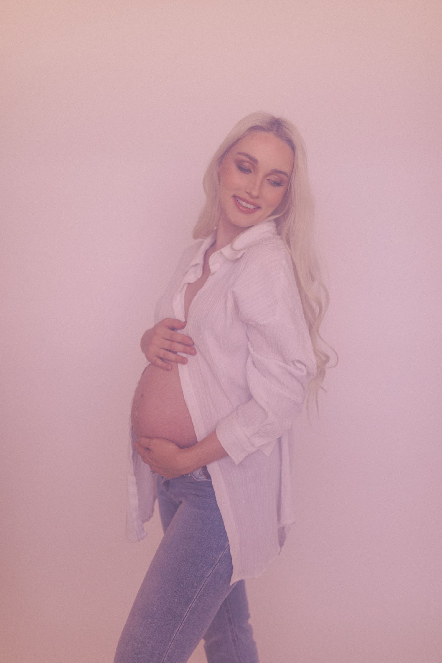 A maternity photoshoot featuring a white button-down shirt and jeans against a pink background.