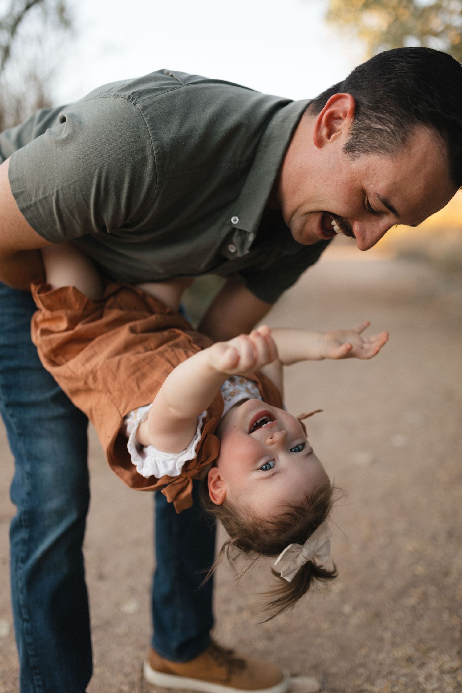 Man in gray sweater dips and embraces a small child in a brown shirt during a playful moment together outdoors.