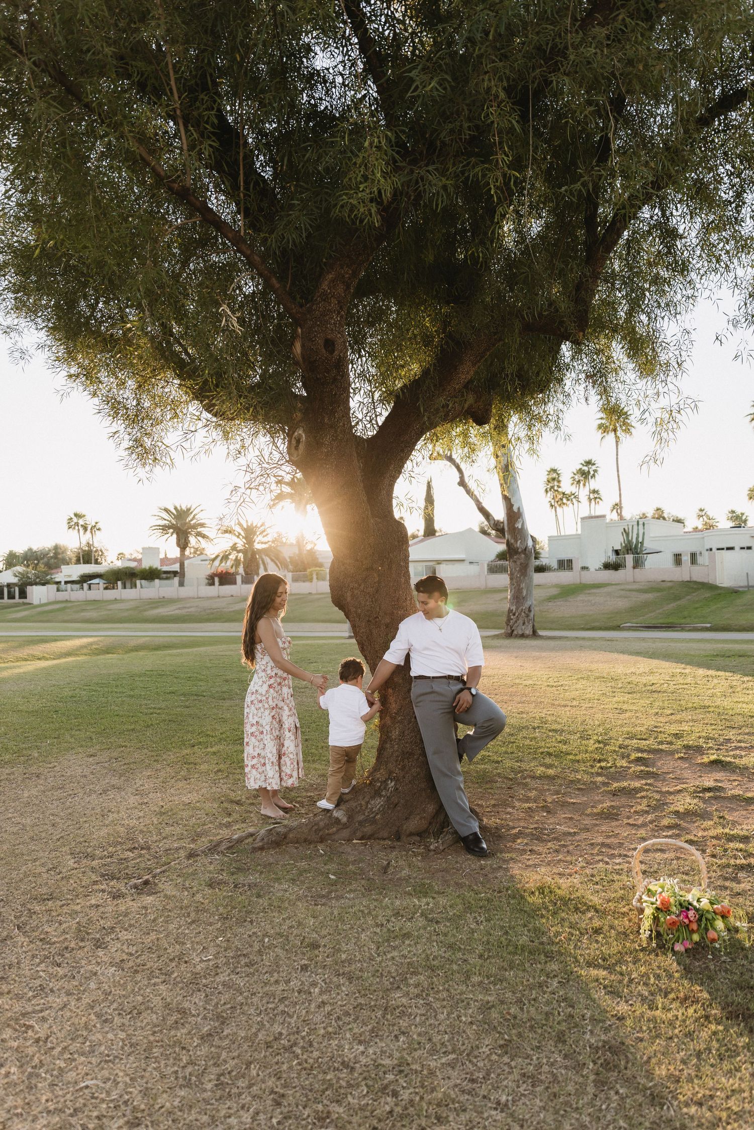 Family of three standing around a large tree at golden hour in a lush green park setting.