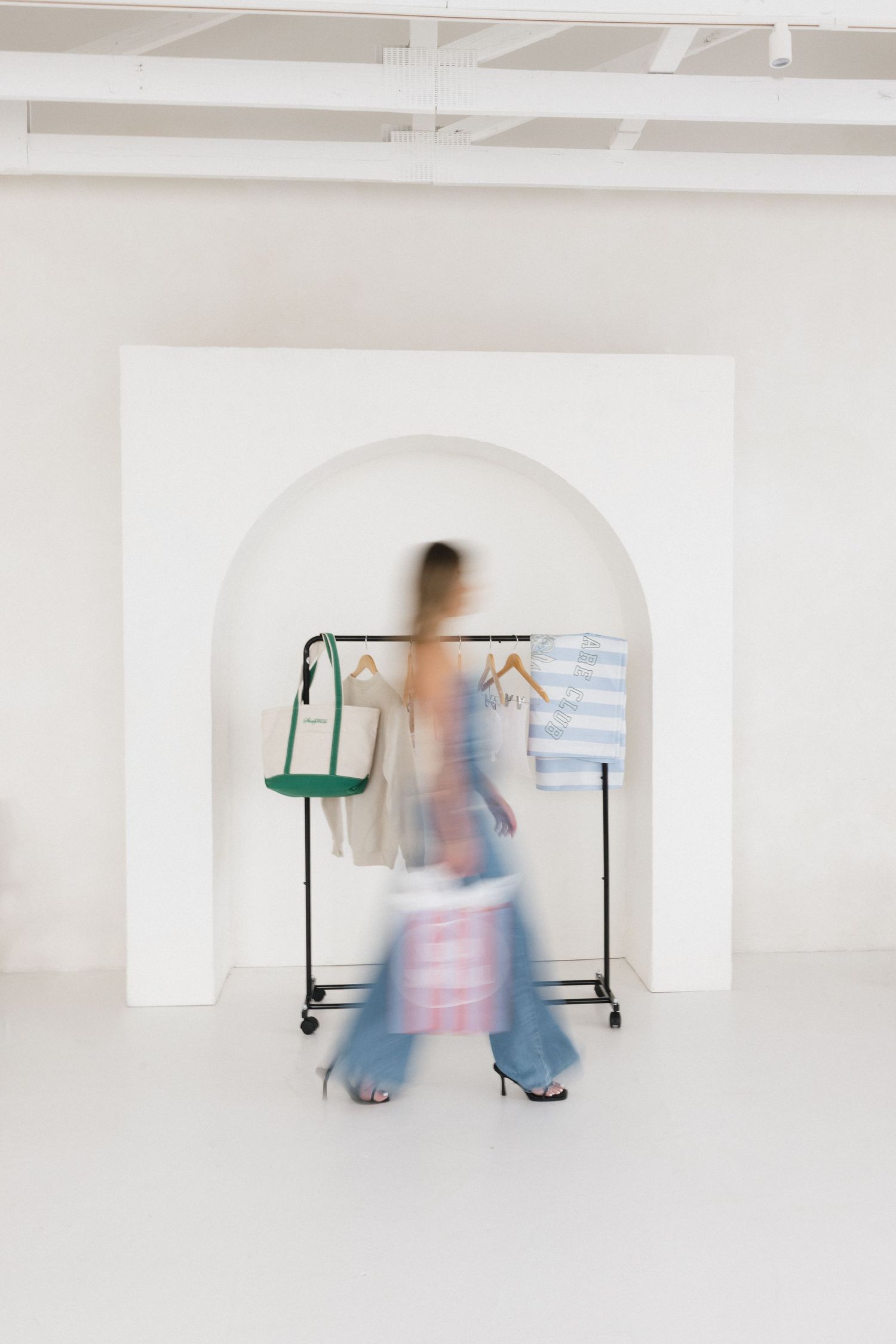 Blurred woman in wide-leg jeans carrying shopping bags near a clothing rack in a minimalist white studio.