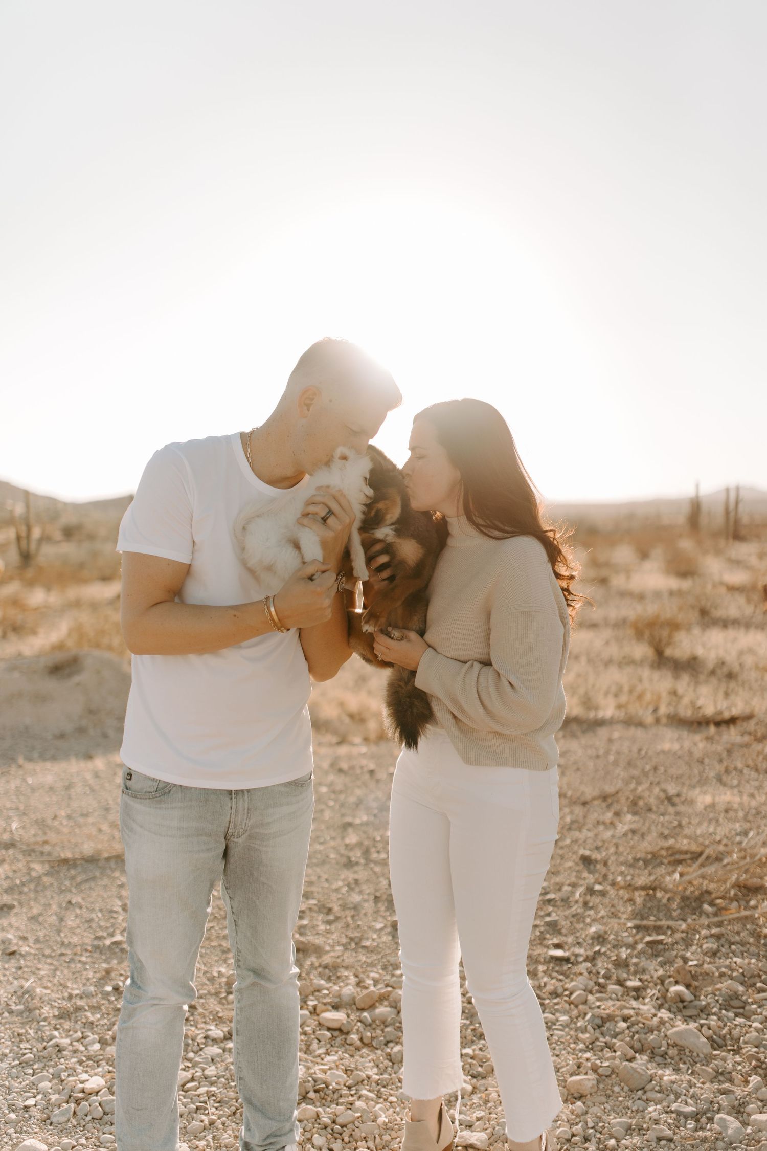 Couple holds their pets while sharing an intimate moment in a desert setting.