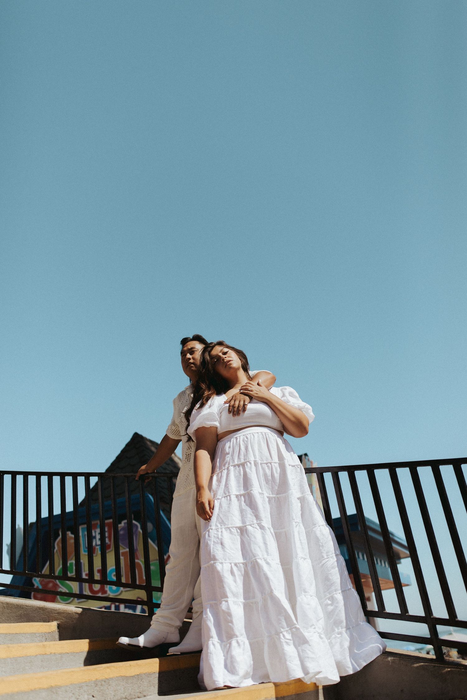 Couple in white clothing poses against blue sky on elevated platform.