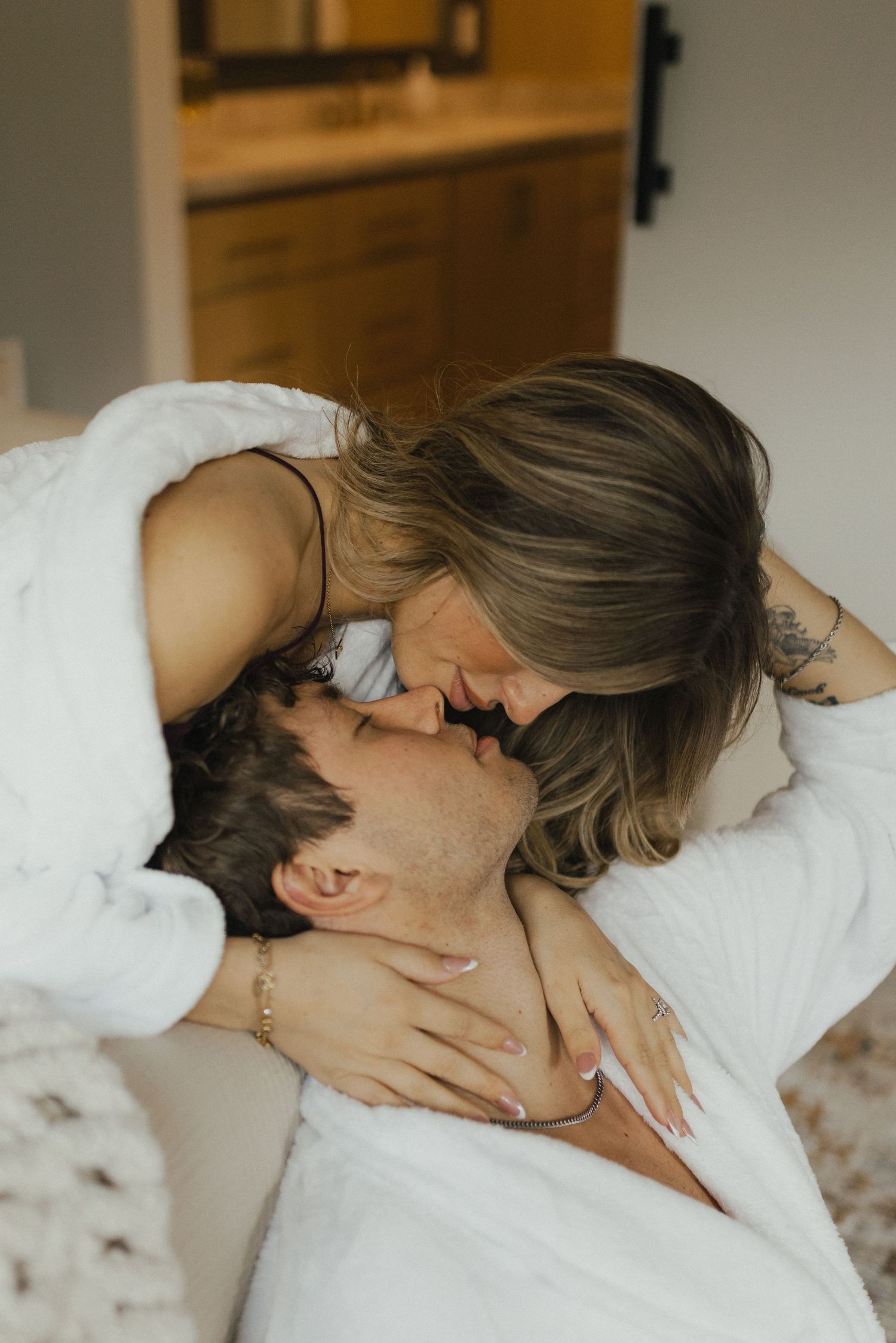 A couple sharing a romantic kiss while wrapped in white blankets on a bed in a cozy bedroom.