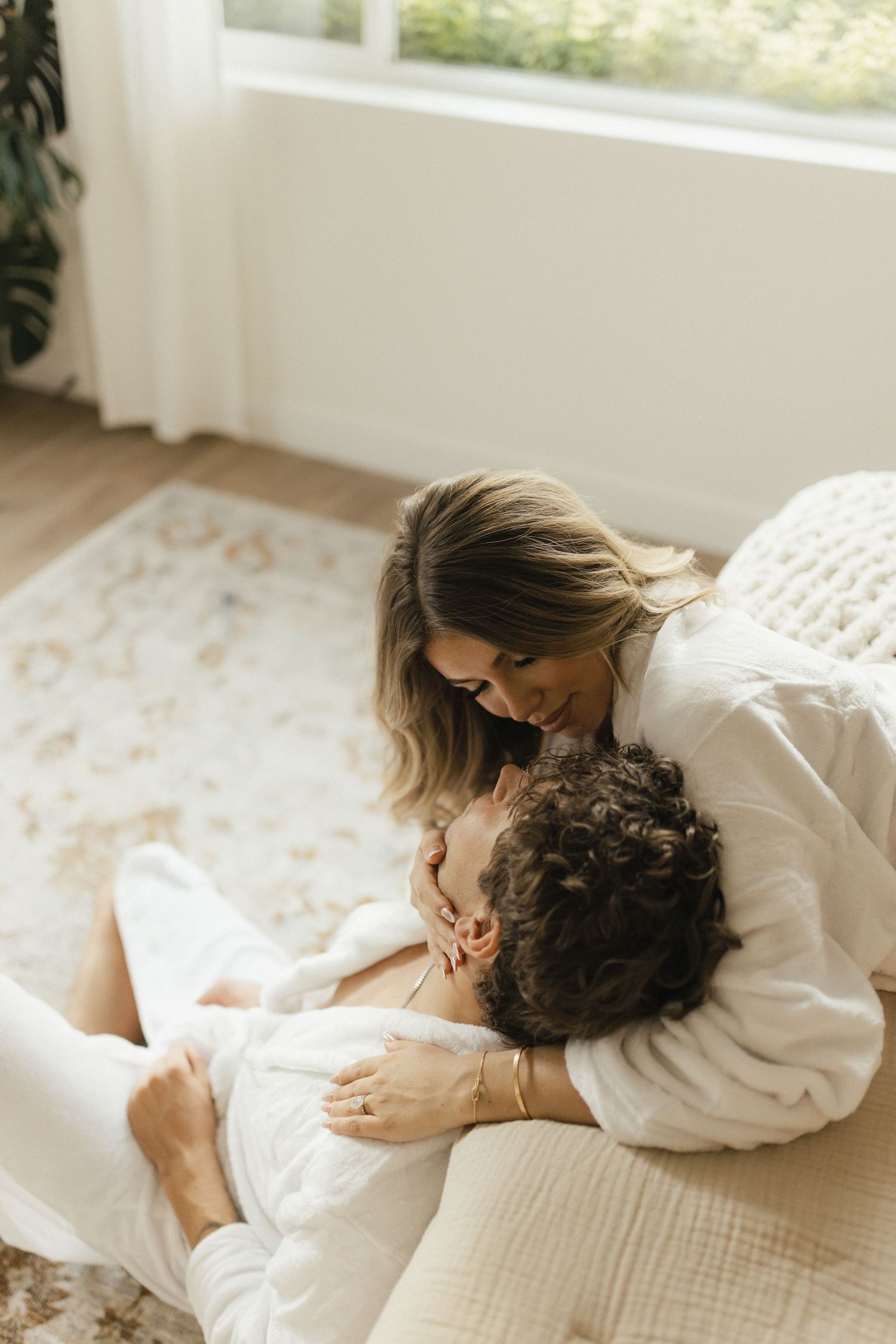Romantic couple in white robes sharing an intimate moment on a cozy bedroom floor near a sunlit window.
