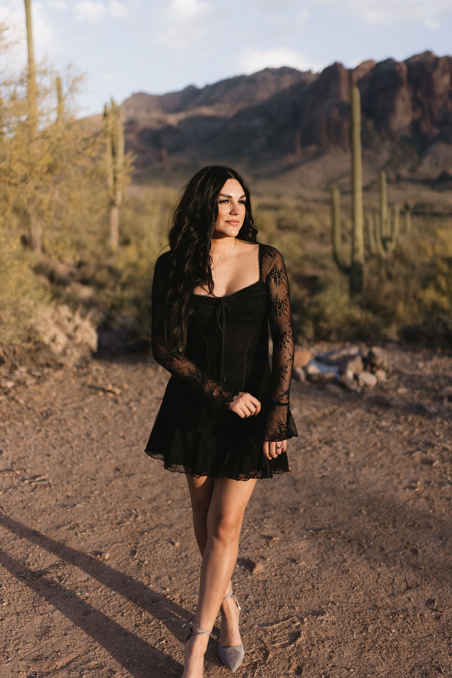 Woman in black lace mini dress poses on desert trail with saguaro cacti and red rock mountains at sunset.