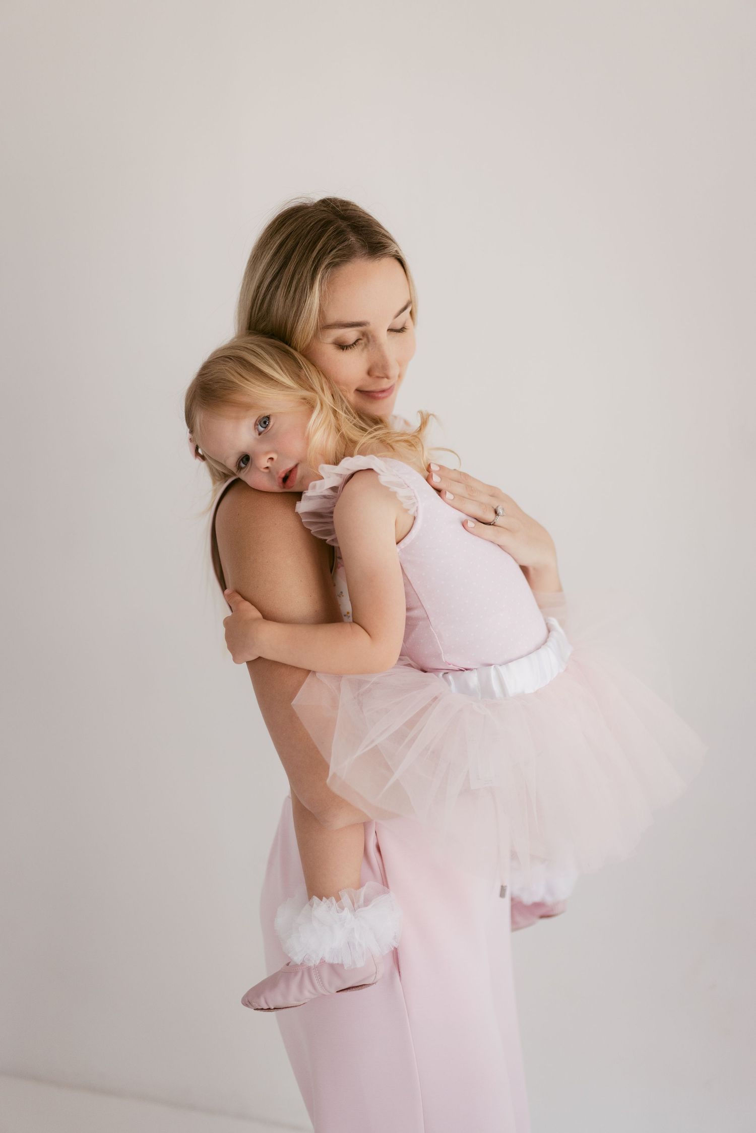 A mother in a pink outfit holds her young daughter in a matching pink tutu dress against a white background.