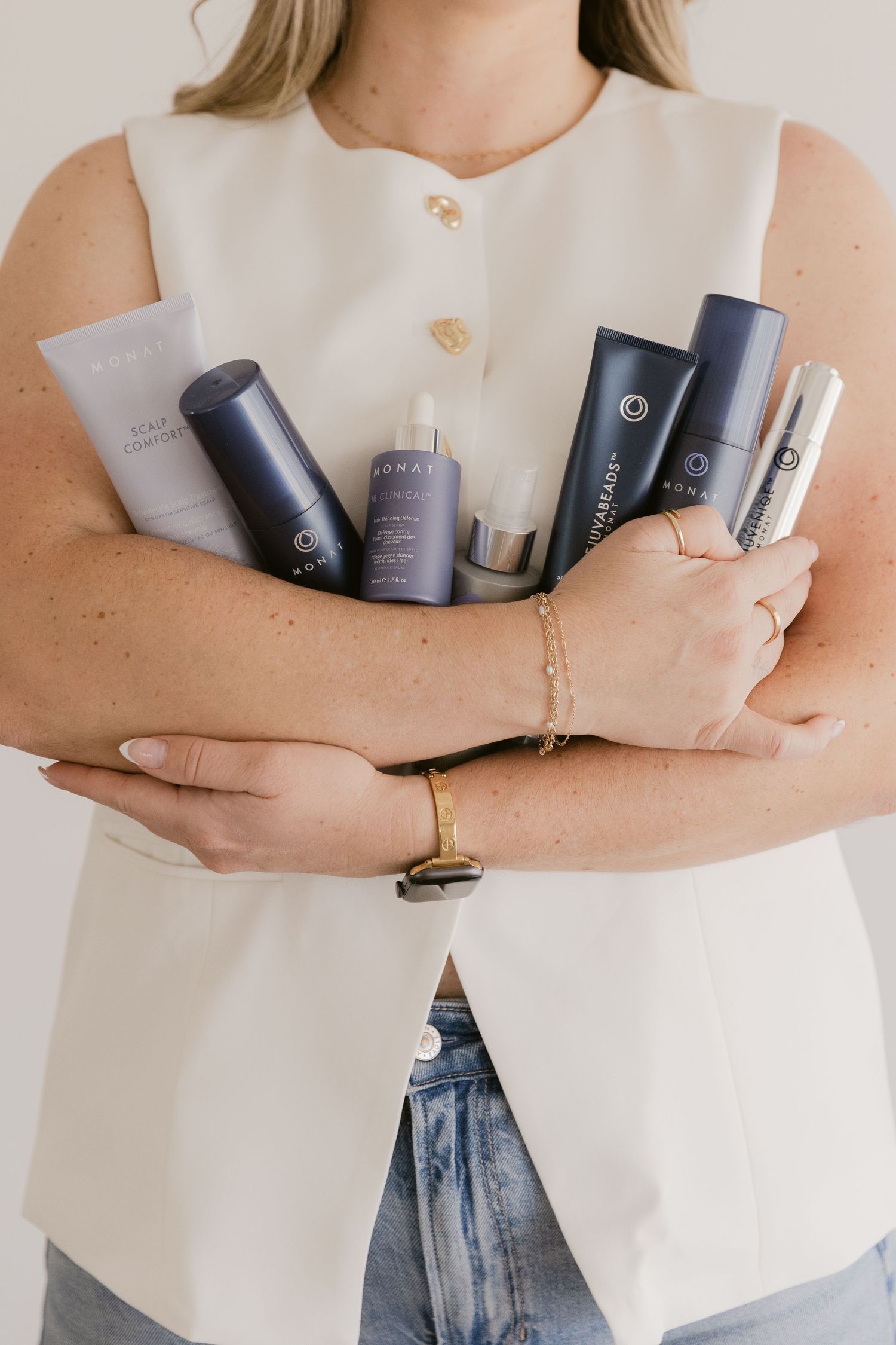 Woman in white sleeveless top holding multiple Monat haircare and skincare products while wearing gold jewelry.
