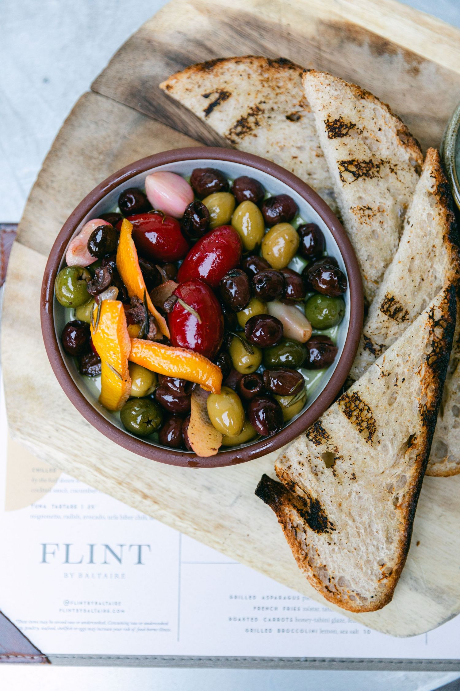 Colorful mixed olives and peppers served in a ceramic bowl with grilled bread on parchment paper.