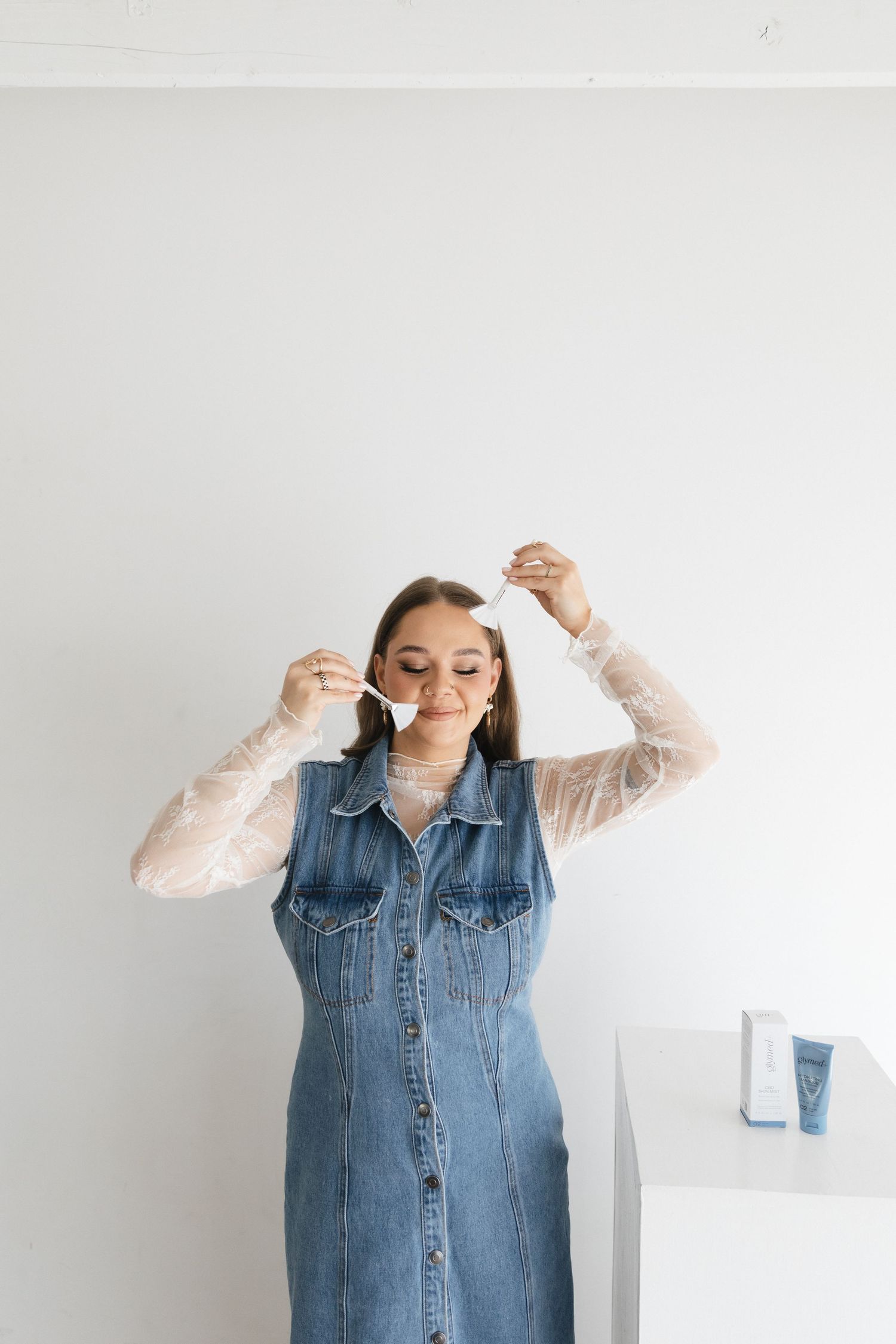 Someone wearing a denim dress and sheer white top holds their hands up playfully against a white wall.