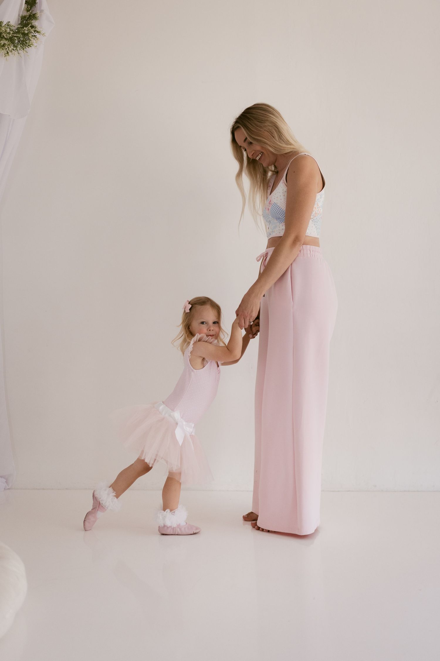 A mother in pink wide-leg pants holds hands with her young daughter in a pink ballet tutu and slippers.