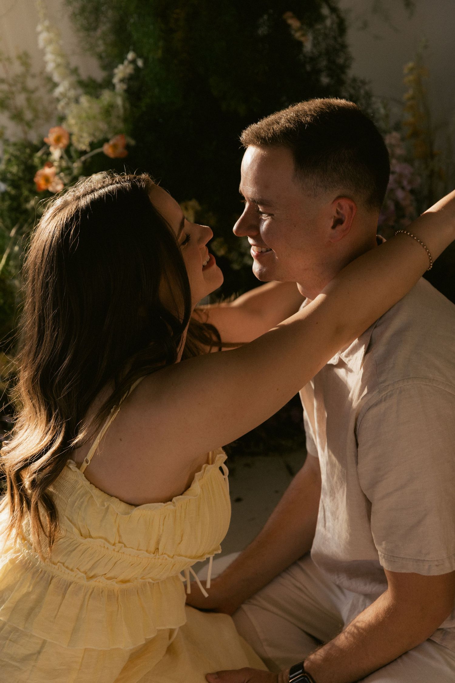A couple shares a romantic moment, woman in yellow dress embracing man in beige shirt, surrounded by lush floral backdrop.