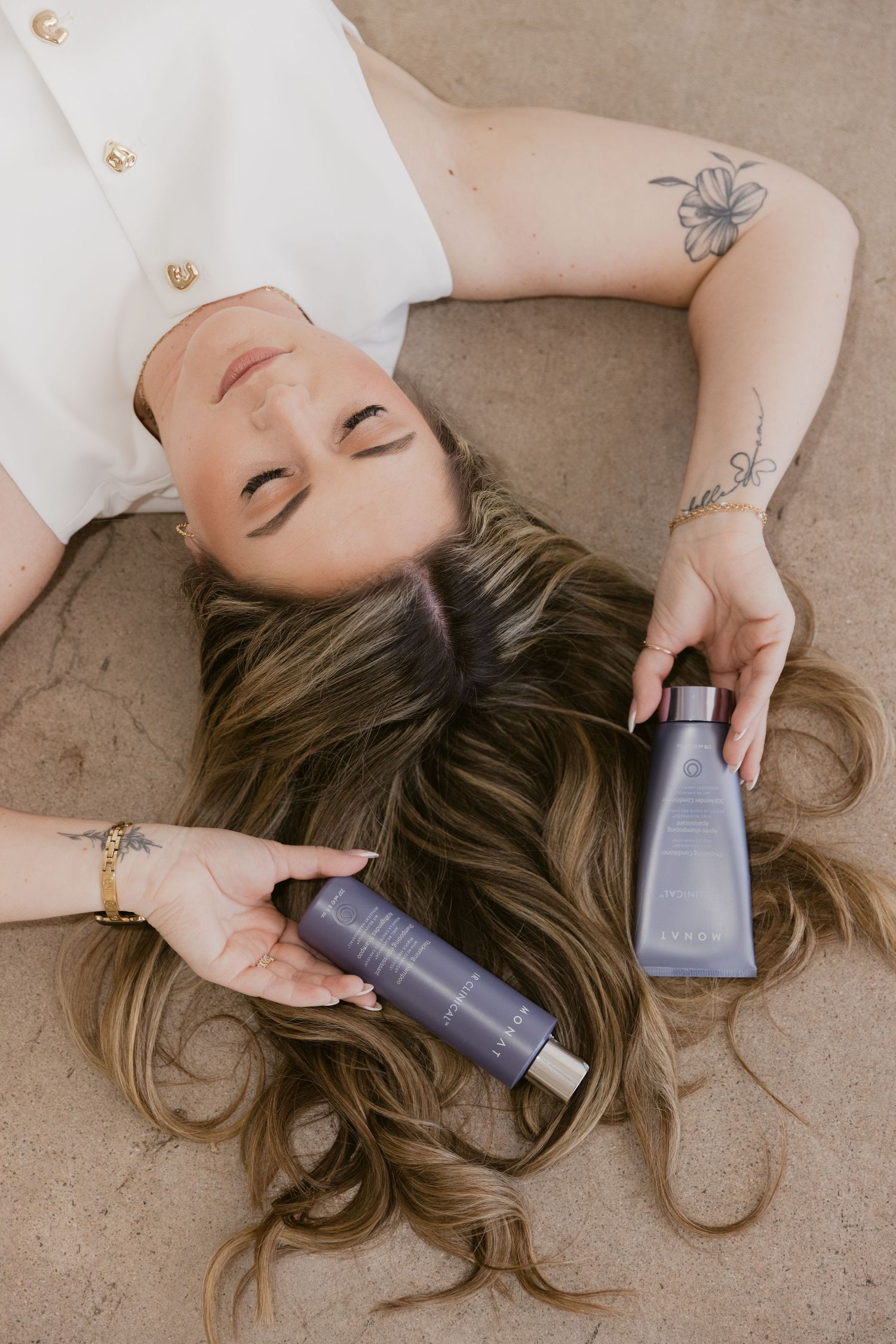 Woman lying down with long wavy hair holding two purple hair care product bottles, showcasing a relaxed beauty lifestyle.