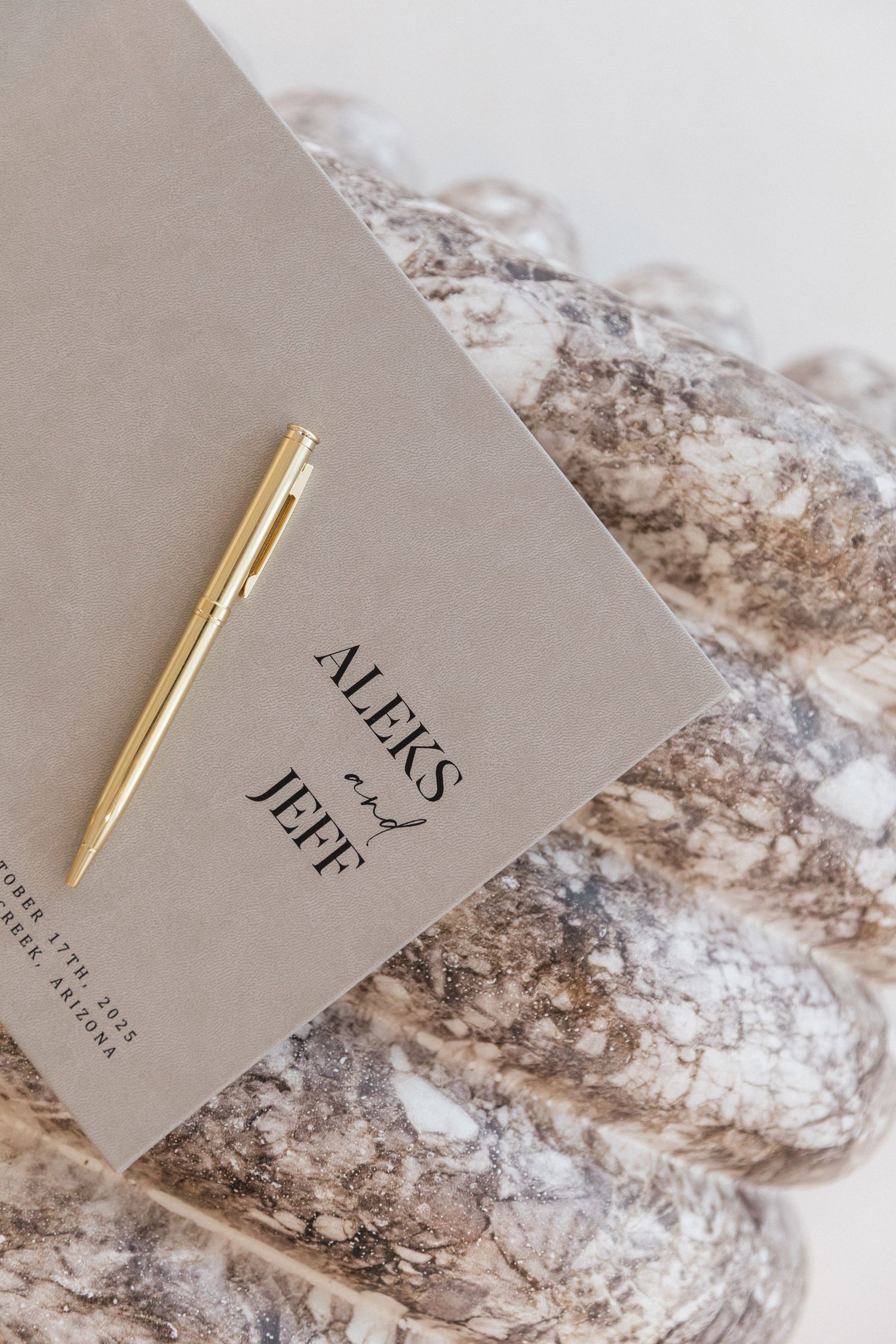 A gold pen rests on a gray notebook with 'Alexis Jeff' text against a marble textured background.