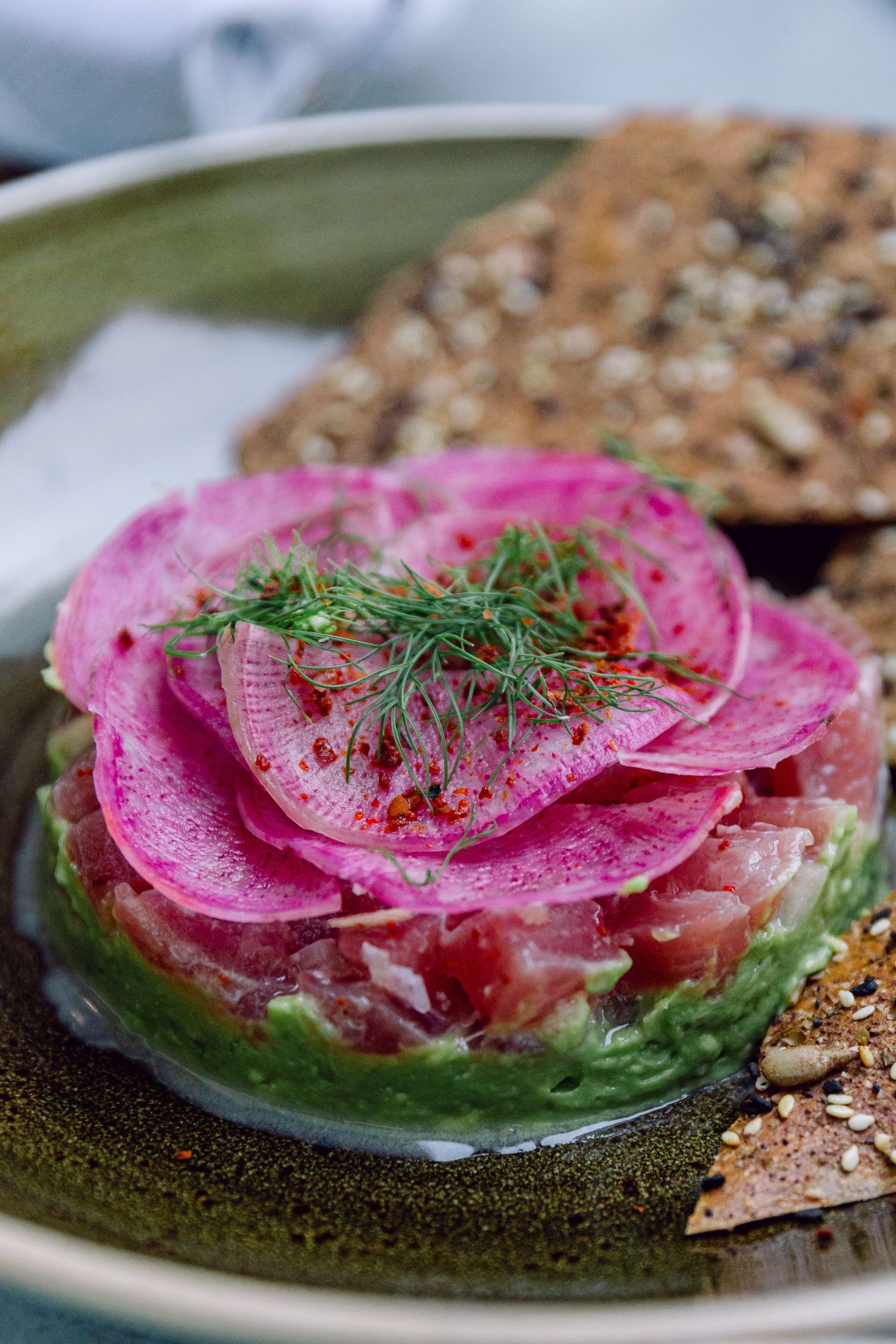 Vibrant pink watermelon radish and dill appetizer served with seeded crackers on a green ceramic plate.