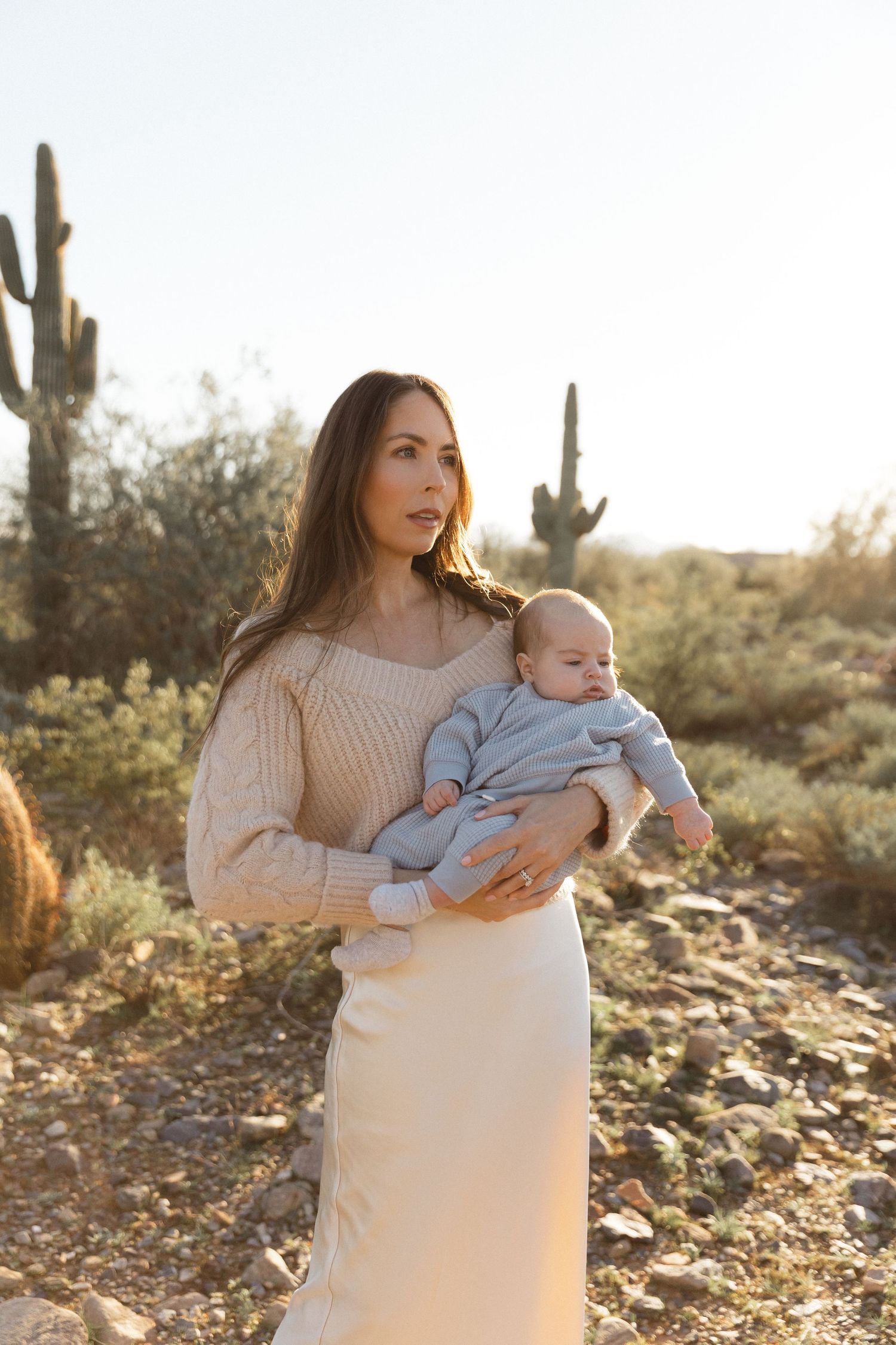 A mother and baby pose in a desert landscape with saguaro cacti at sunset wearing cream colored clothing.