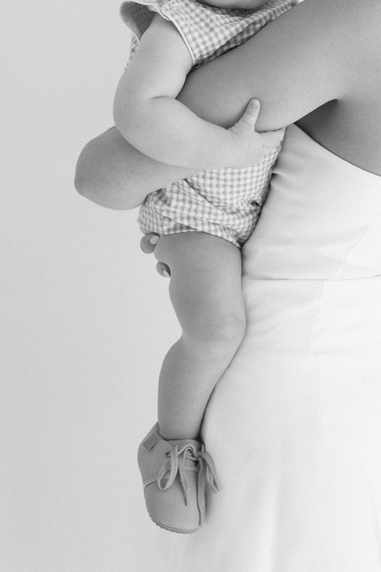 Black and white photo of a mother holding a baby in gingham outfit and soft shoes, capturing a tender moment.