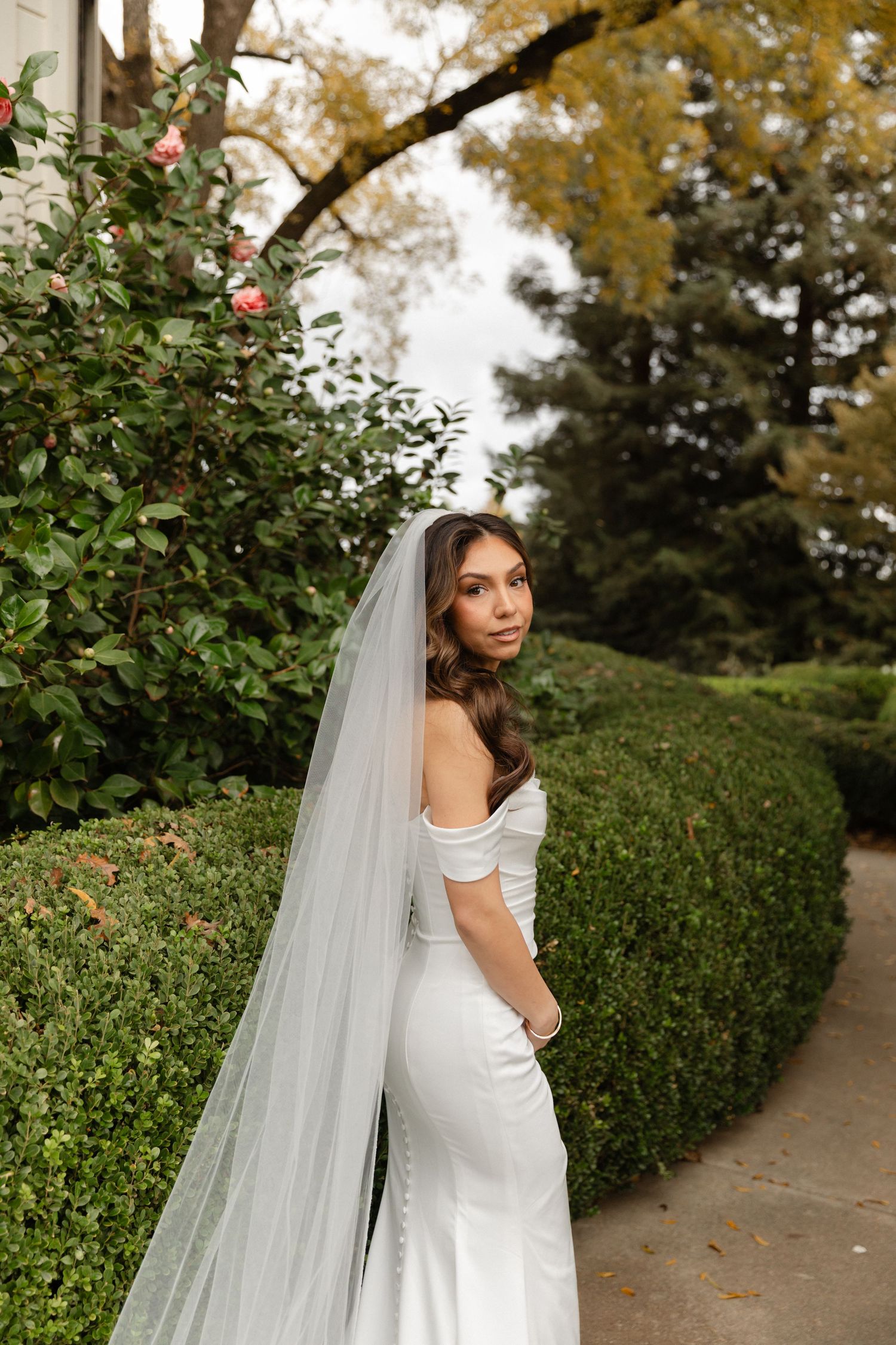 A bride in a white dress and flowing veil poses gracefully in a garden path lined with green hedges and autumn trees.