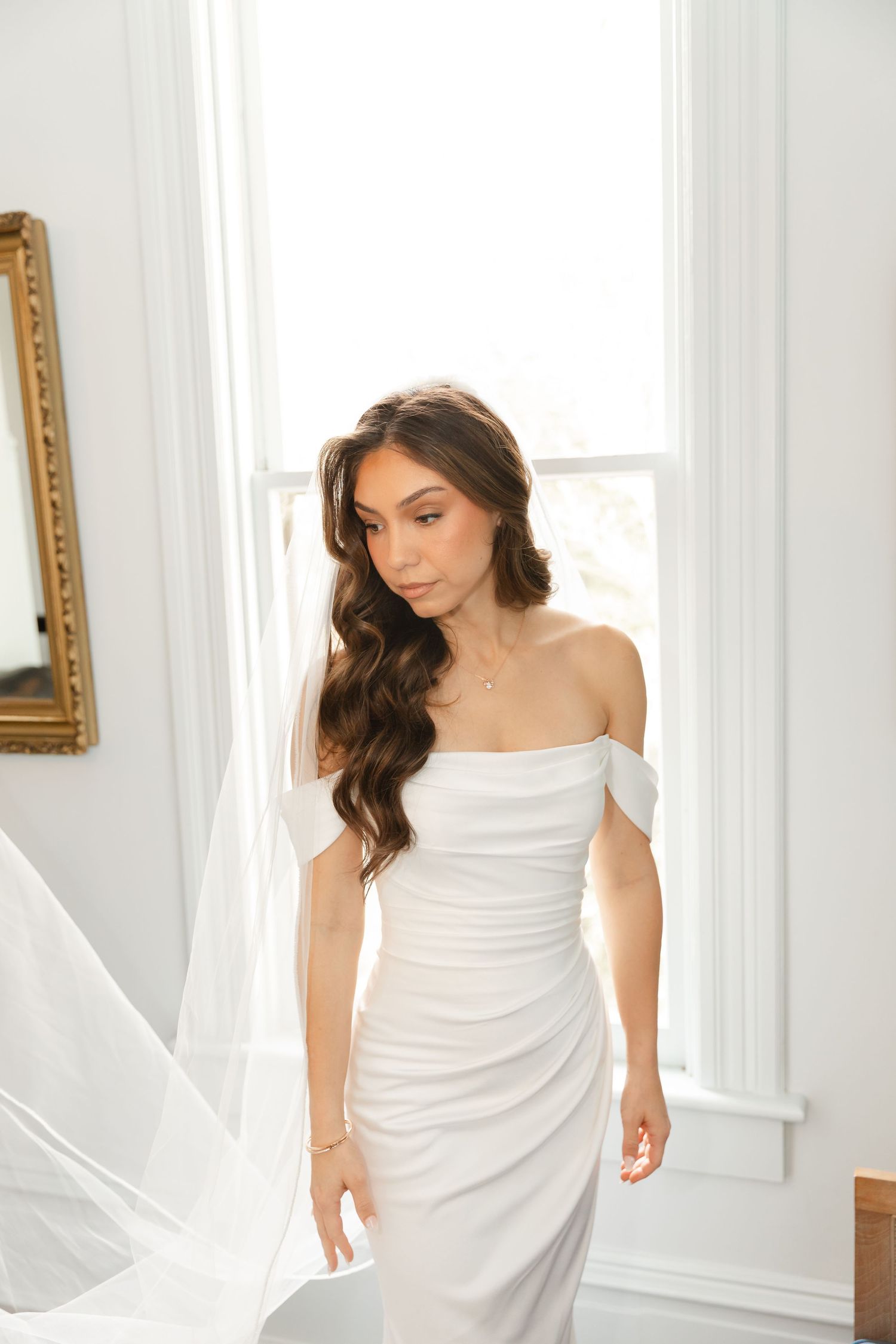 A woman in a white draped dress poses elegantly against a bright white interior with sunlit windows.