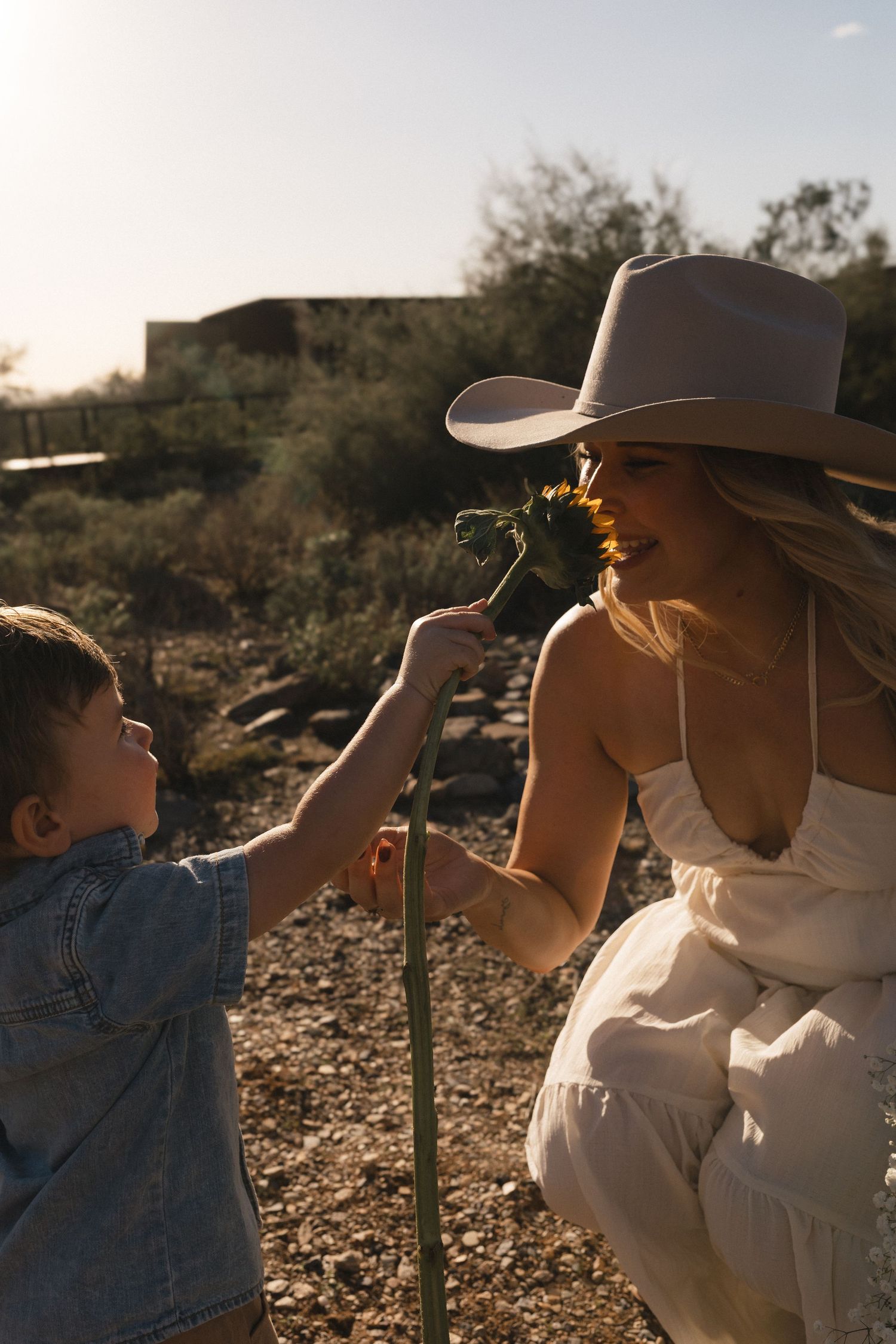 A tender moment shared in the desert as a child and woman in a hat examine a sunflower at sunset.