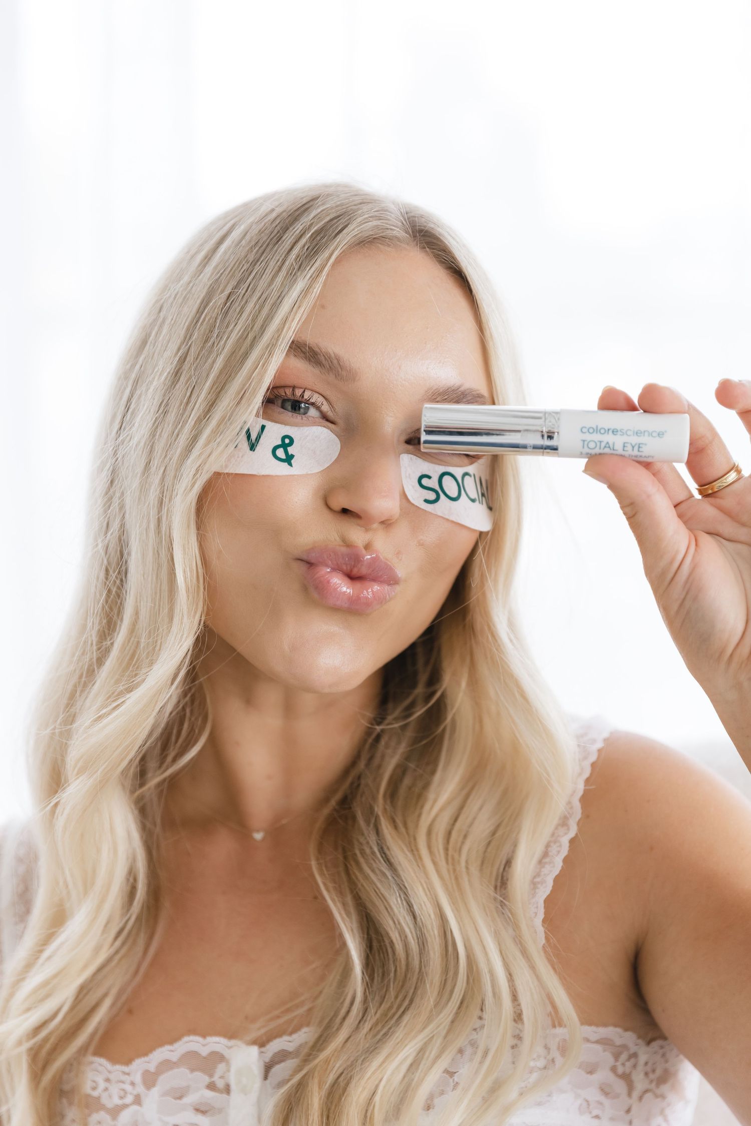 Blonde woman wearing eye patches holding a skincare product, posing with a pout against a white background.