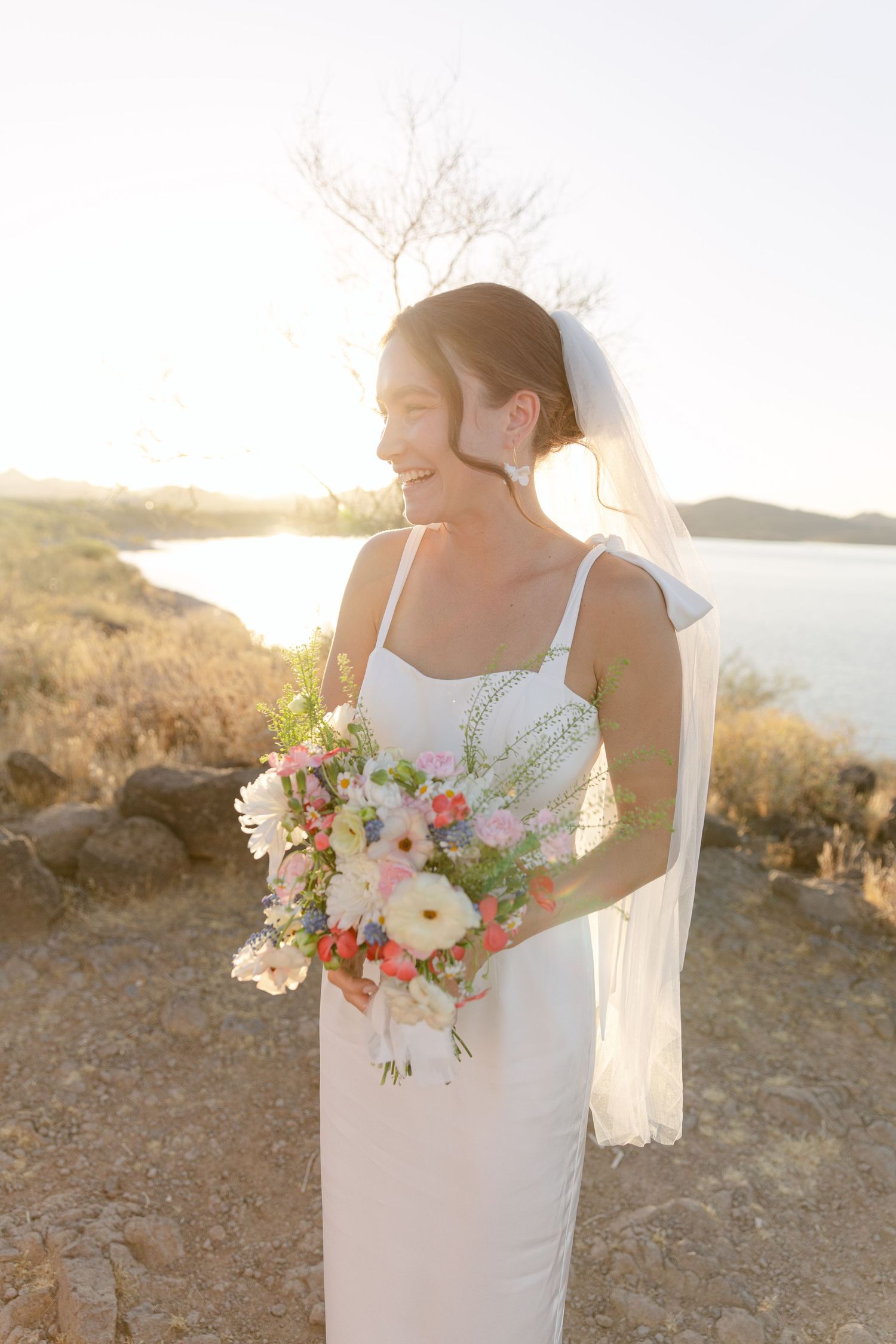 A radiant bride in white holding a colorful bouquet against a sunset lake view.