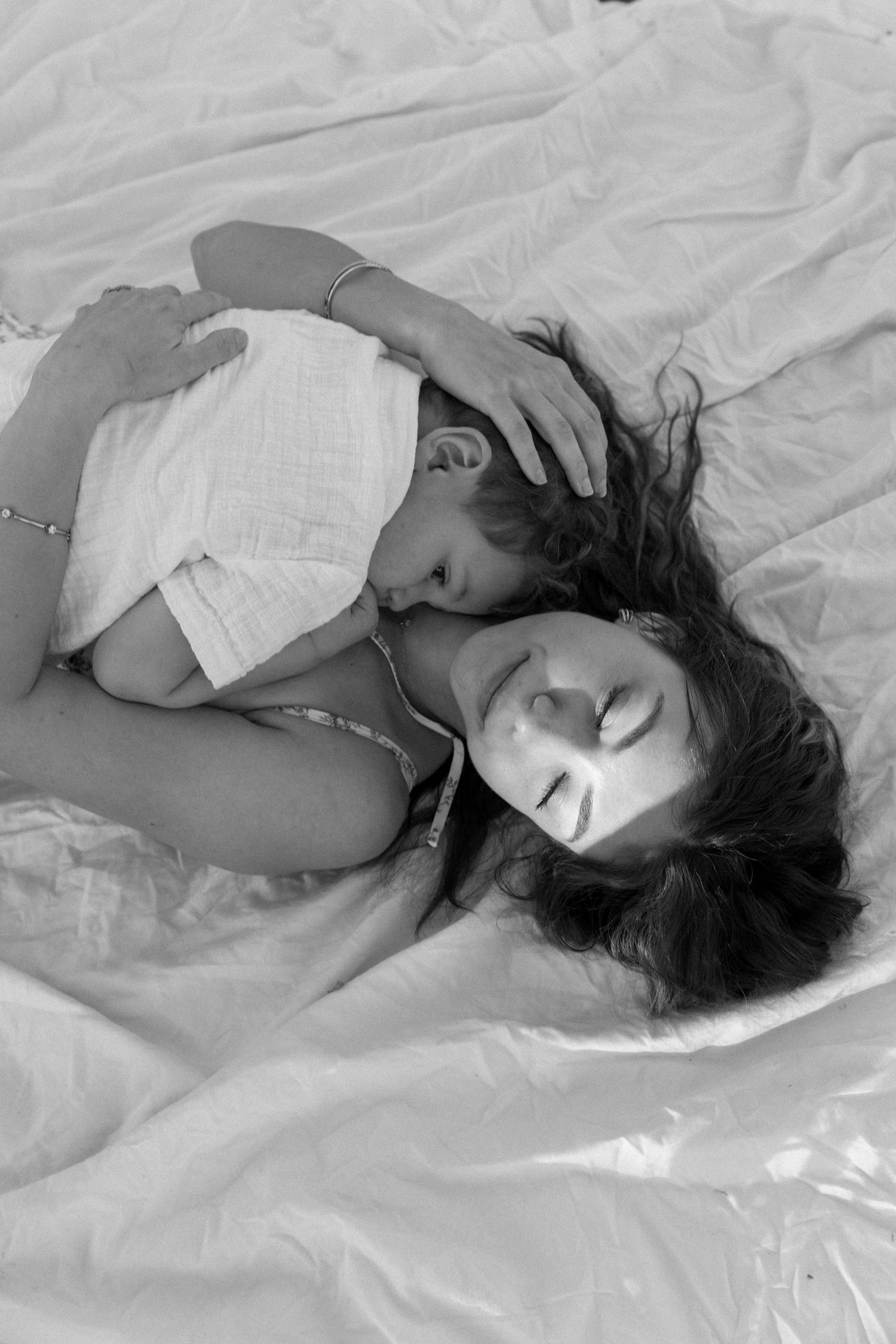 Black and white photo of a mother lying on white bedding while her young child cuddles and kisses her cheek tenderly.
