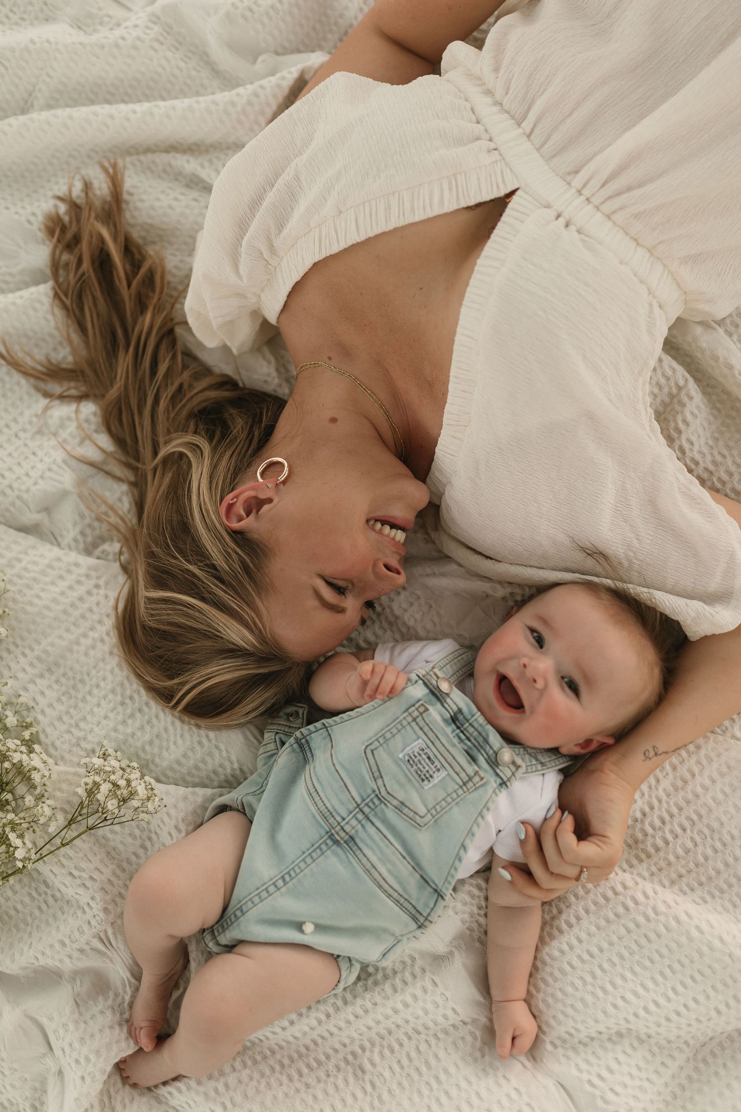 A child lying on a white textured blanket wearing a mint green romper while smiling and playing in a natural light setting.