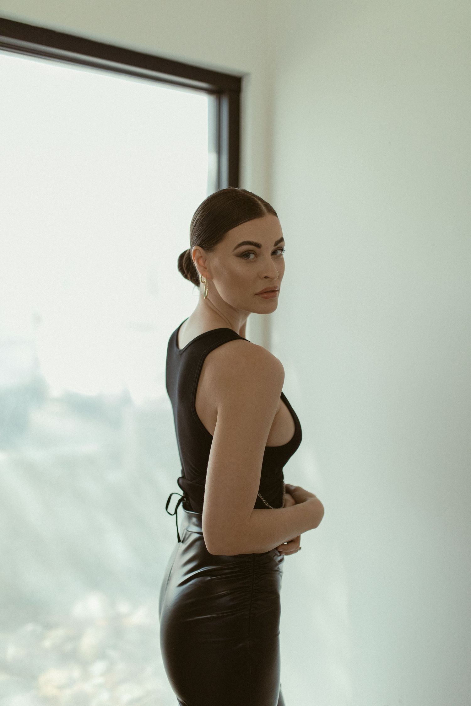 Fashion model in black dress poses against white wall with dramatic lighting.