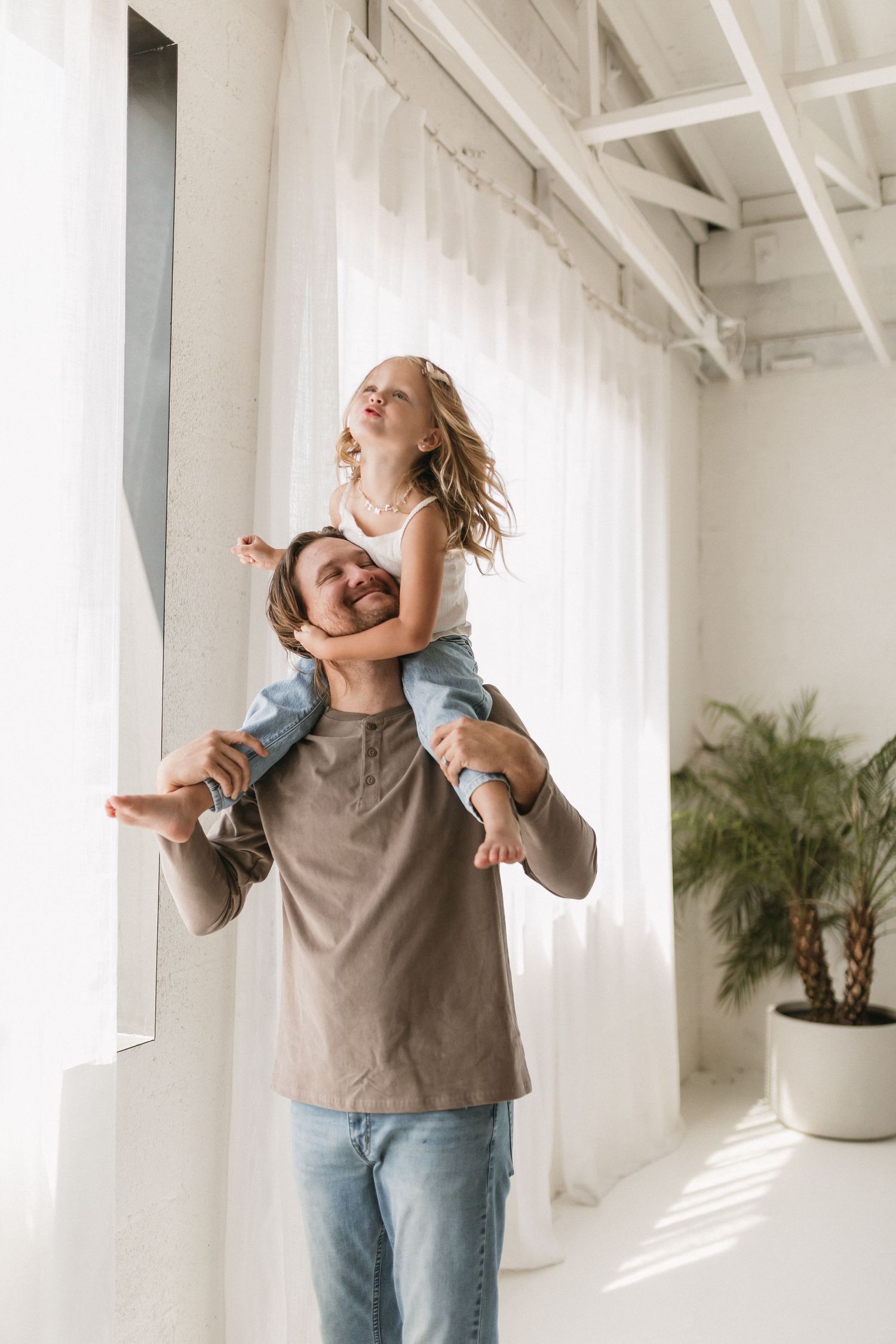 Parent and child embrace joyfully in a bright white room with natural light and potted plants.
