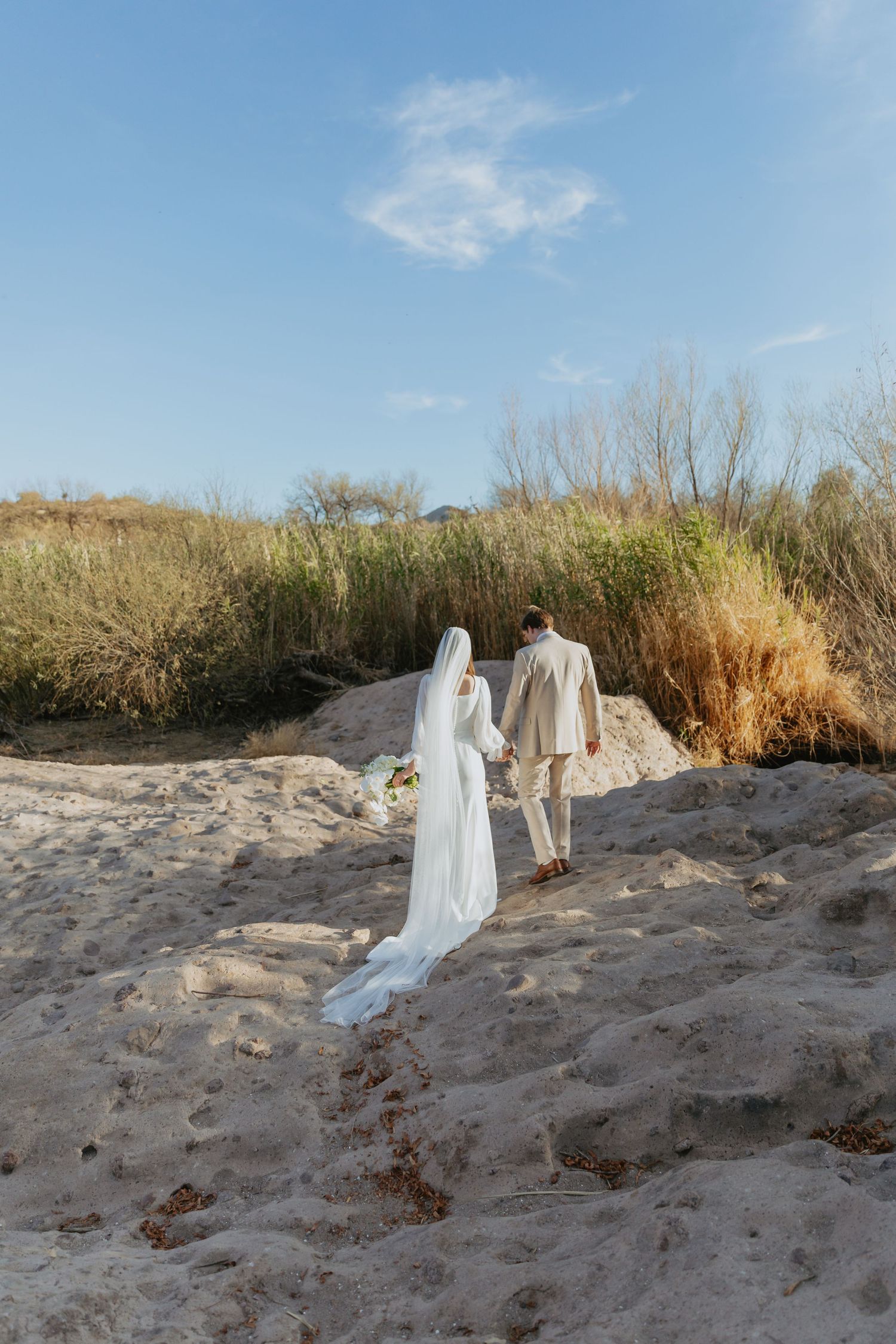 Two figures in flowing white dresses walk along a sandy beach path with tall grass and blue sky in the background.